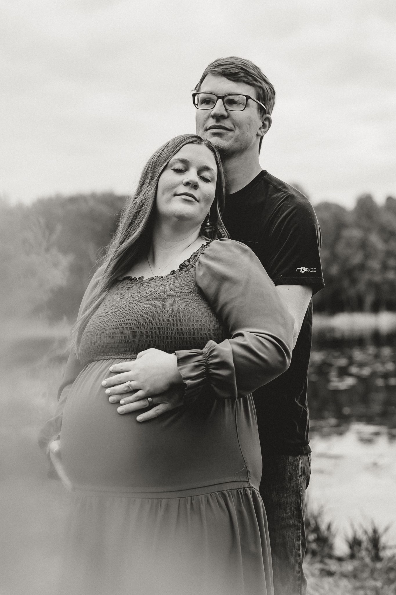 Black and white maternity photo of a pregnant woman and her partner embracing near a misty lake.