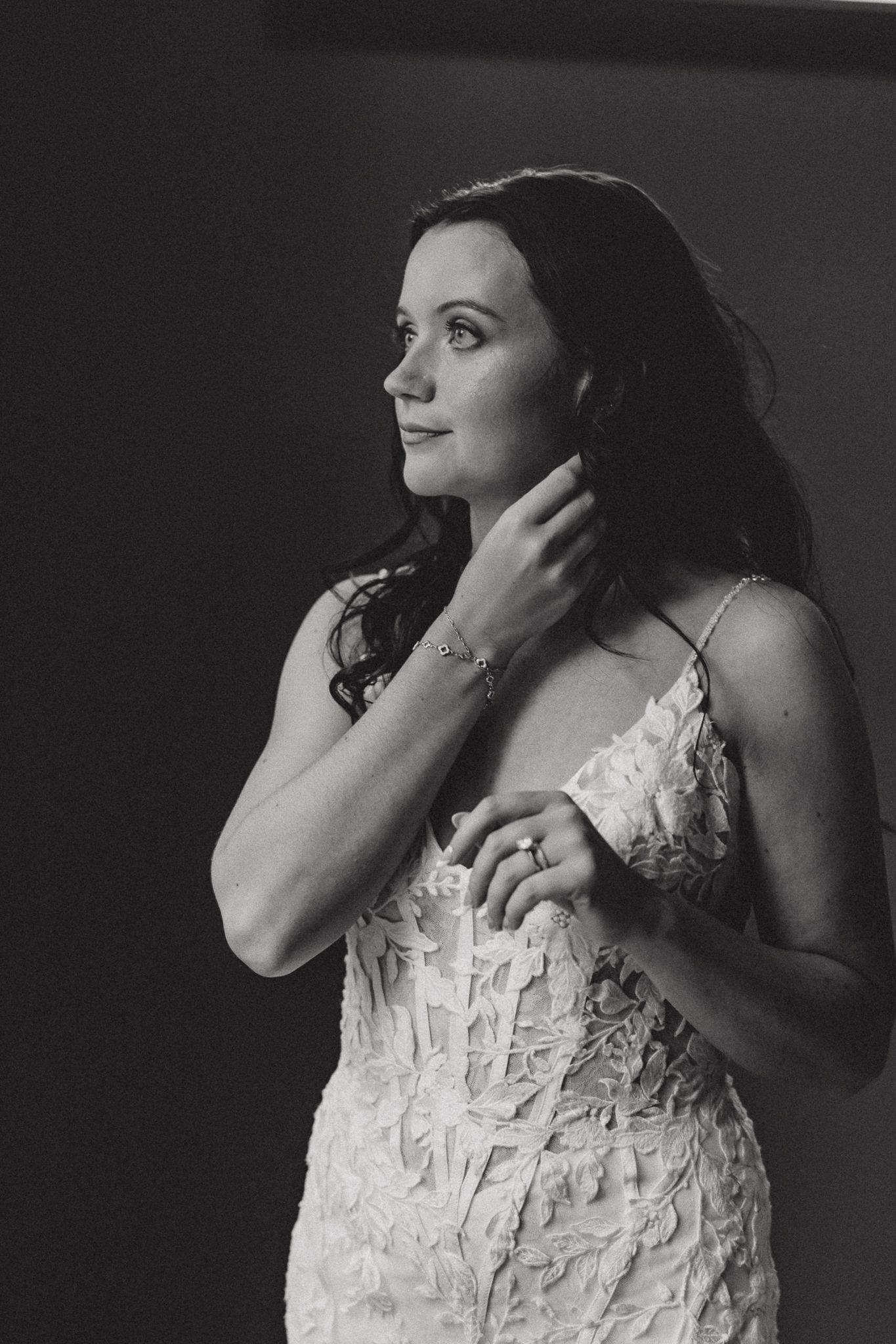 Black and white portrait of a bride in a lace wedding gown touching her hair and gazing thoughtfully to the side.