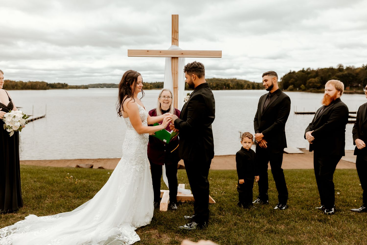 Bride in a white lace gown and groom hold hands during an outdoor lakeside wedding ceremony beneath a wooden cross as guests look on.