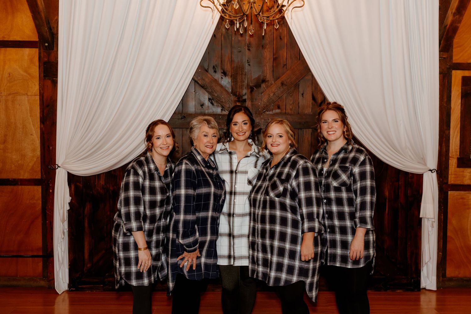 Five women in matching black and white plaid flannel shirts pose together in a rustic barn with white draped curtains and a chandelier.