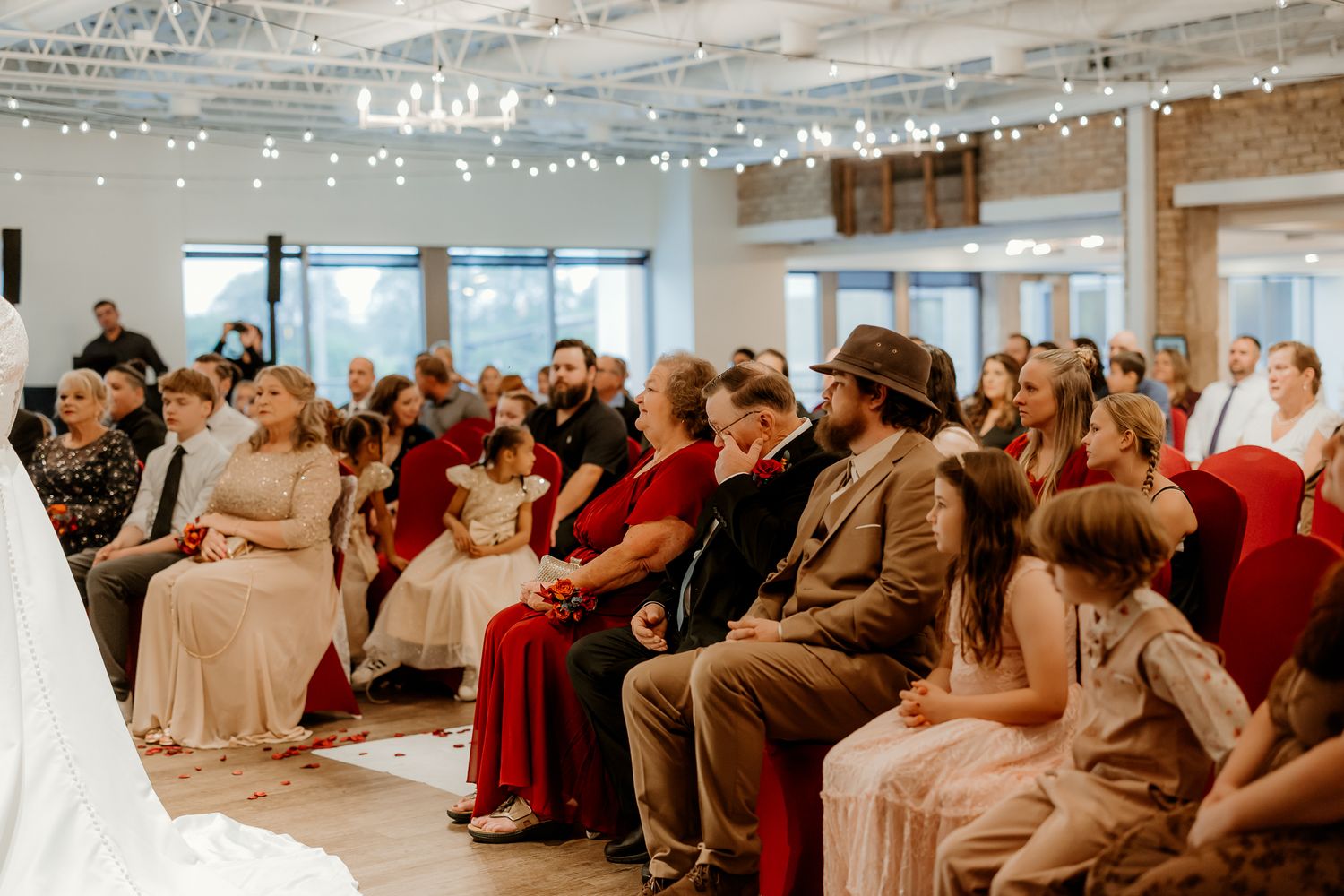 Wedding guests in formal attire sit in rows of chairs watching a ceremony inside a bright venue with string lights and large windows.
