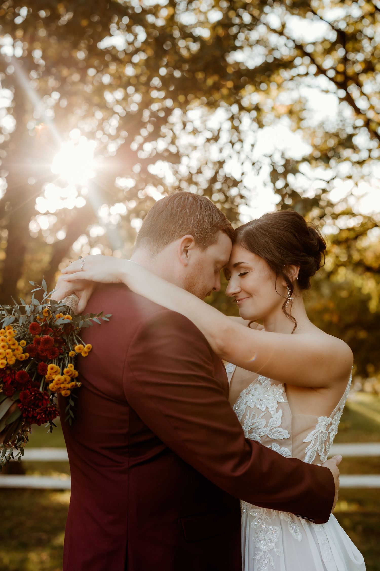 A bride in a lace gown and groom in a burgundy suit embrace forehead to forehead in golden sunset light, holding an autumn bouquet.