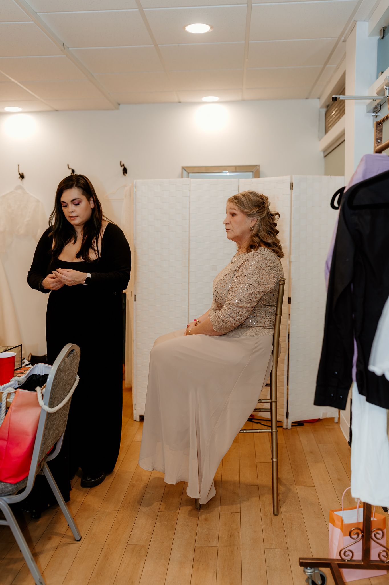 Woman in a beige gown and lace top sits on a chair while a woman in black stands nearby in a bright bridal dressing room.