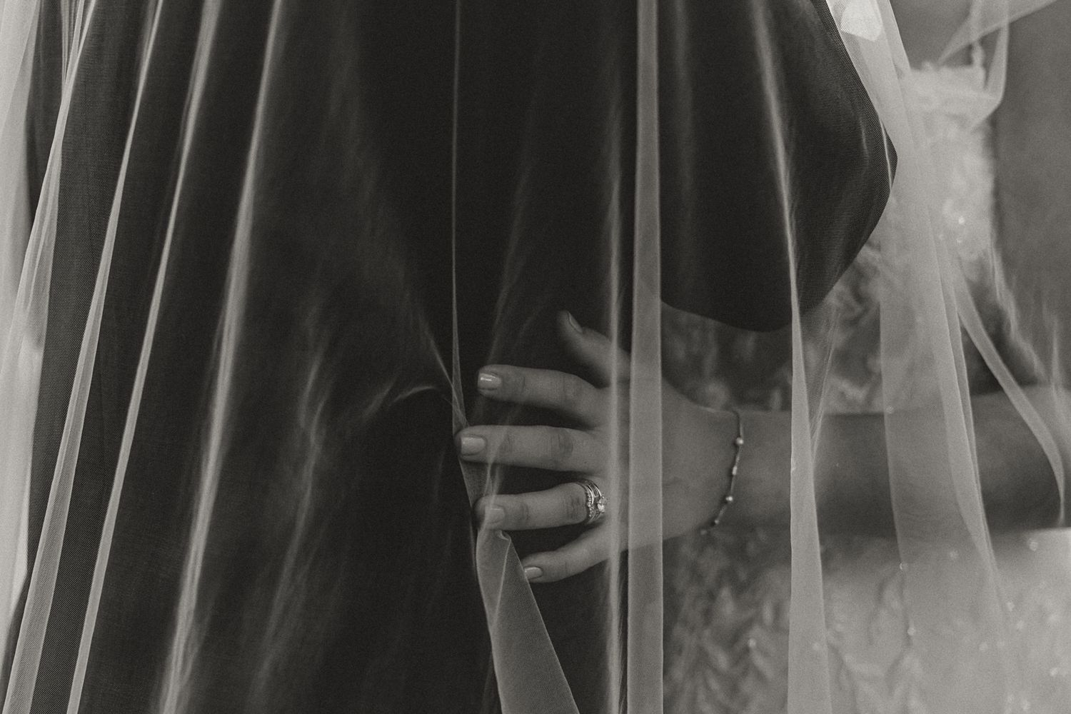 Close-up black and white photo of a bride holding her wedding dress with a delicate ring on her finger.