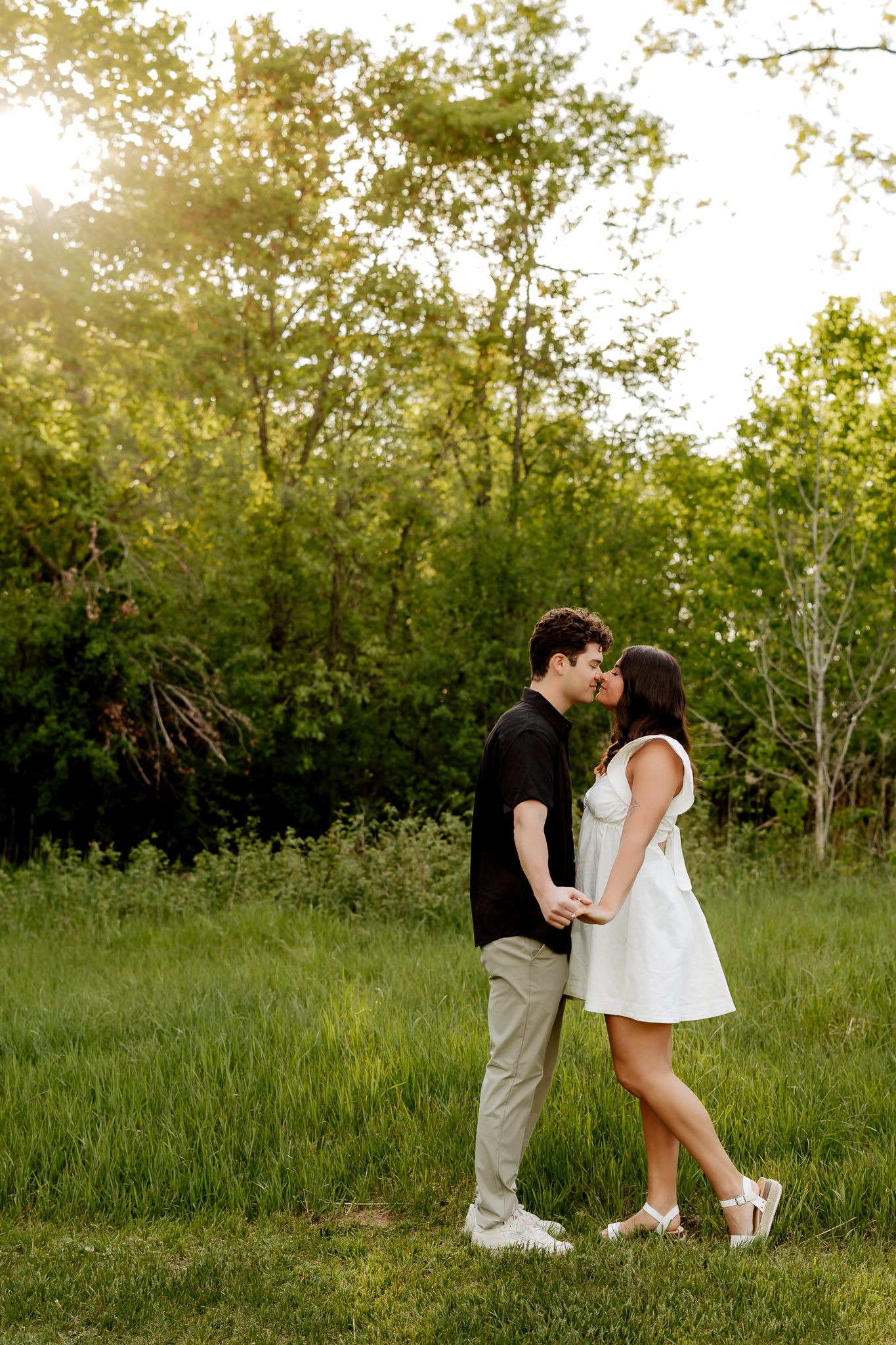 A couple leans in for a kiss while holding hands in a sunlit green meadow surrounded by trees.