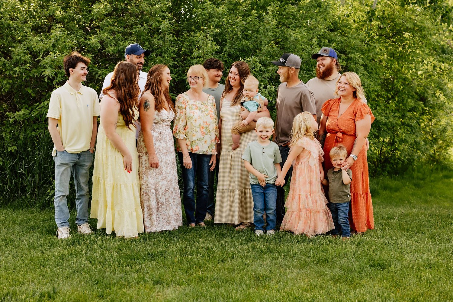 Large family group portrait outdoors on green grass surrounded by lush trees in warm summer light.