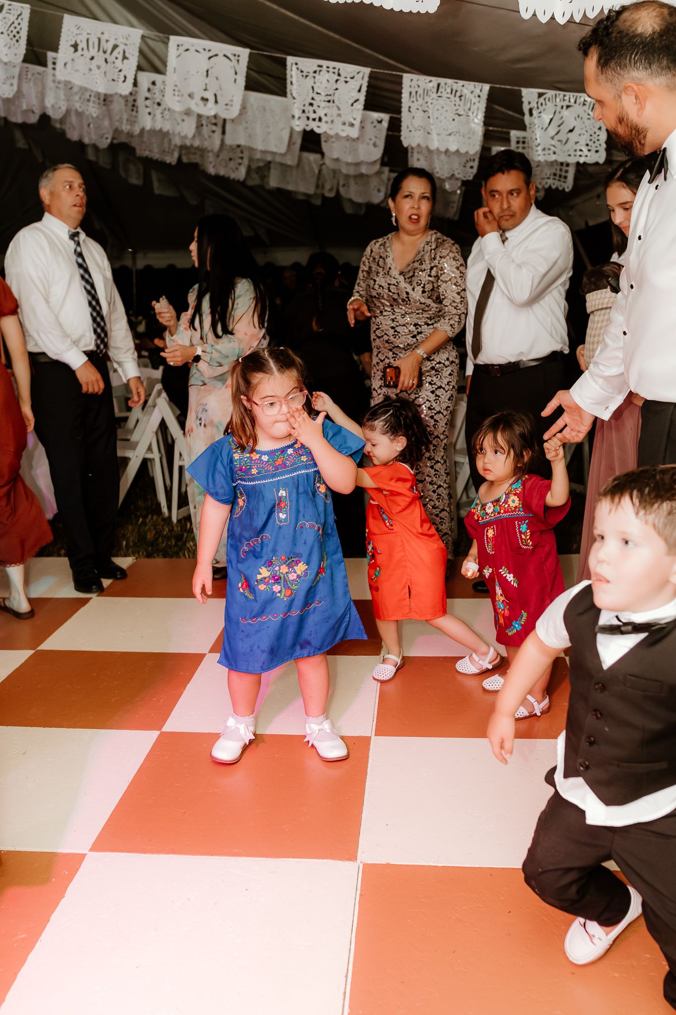 Children dancing on a checkered dance floor at a festive celebration with papel picado decorations overhead.