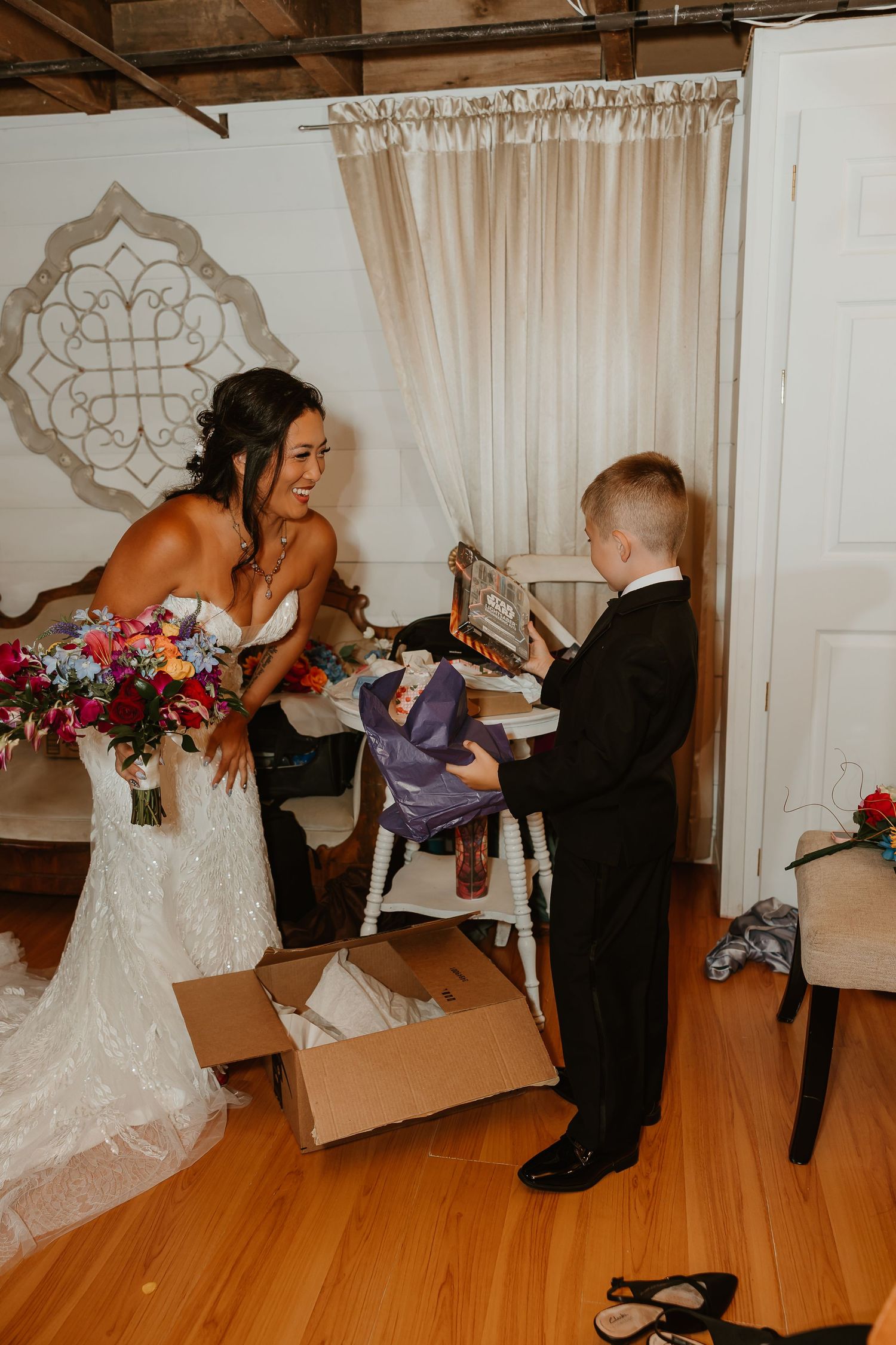 Bride in lace gown laughs with young boy in tuxedo opening a gift box at a wedding venue.