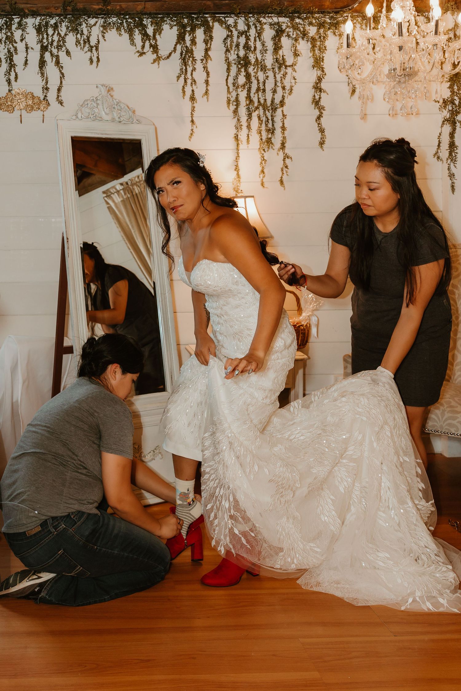 Bride in white lace wedding gown with red heels being assisted by two helpers in a decorated bridal suite.