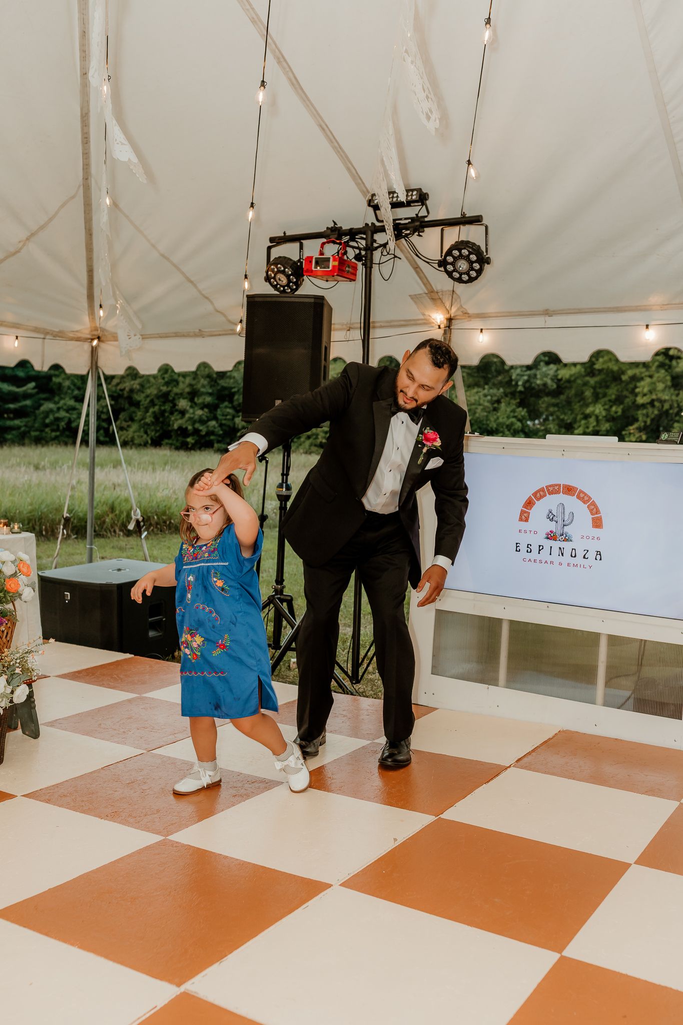 Groom in black suit dancing with young girl in blue dress on checkered dance floor at outdoor wedding reception under tent.