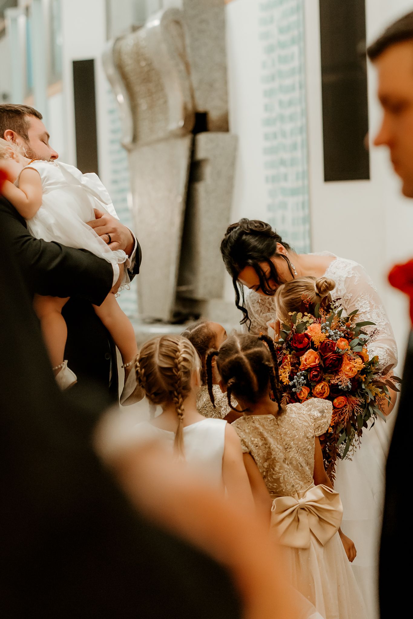 A bride in a lace gown holds an orange and red bouquet while flower girls in braids look on and a man in a suit carries a child.