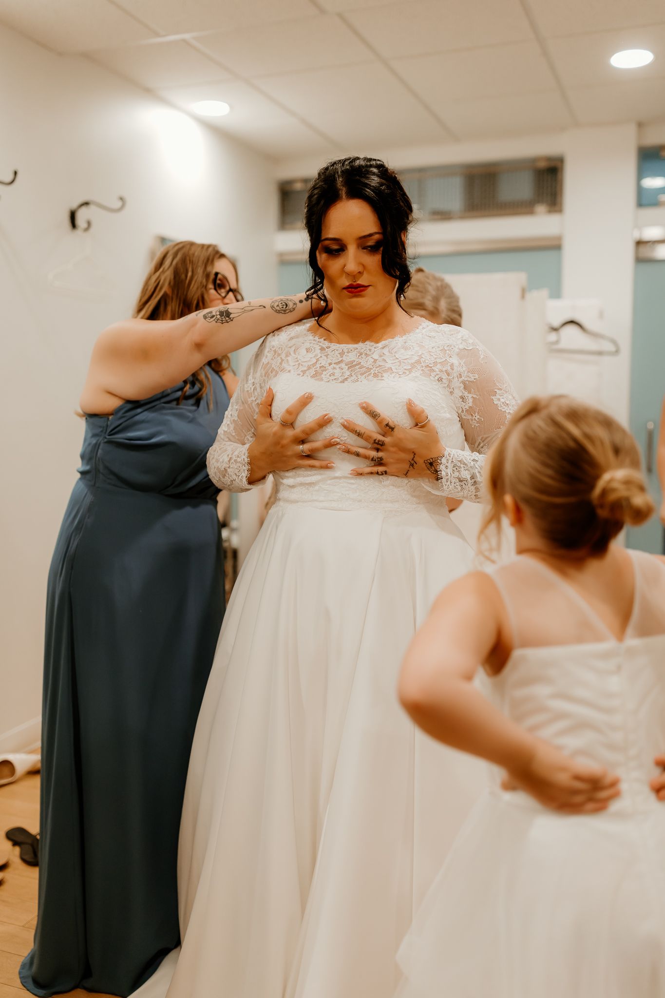 A bride in a lace-sleeved wedding gown is helped into her dress by a bridesmaid in blue and a flower girl in a getting-ready room.