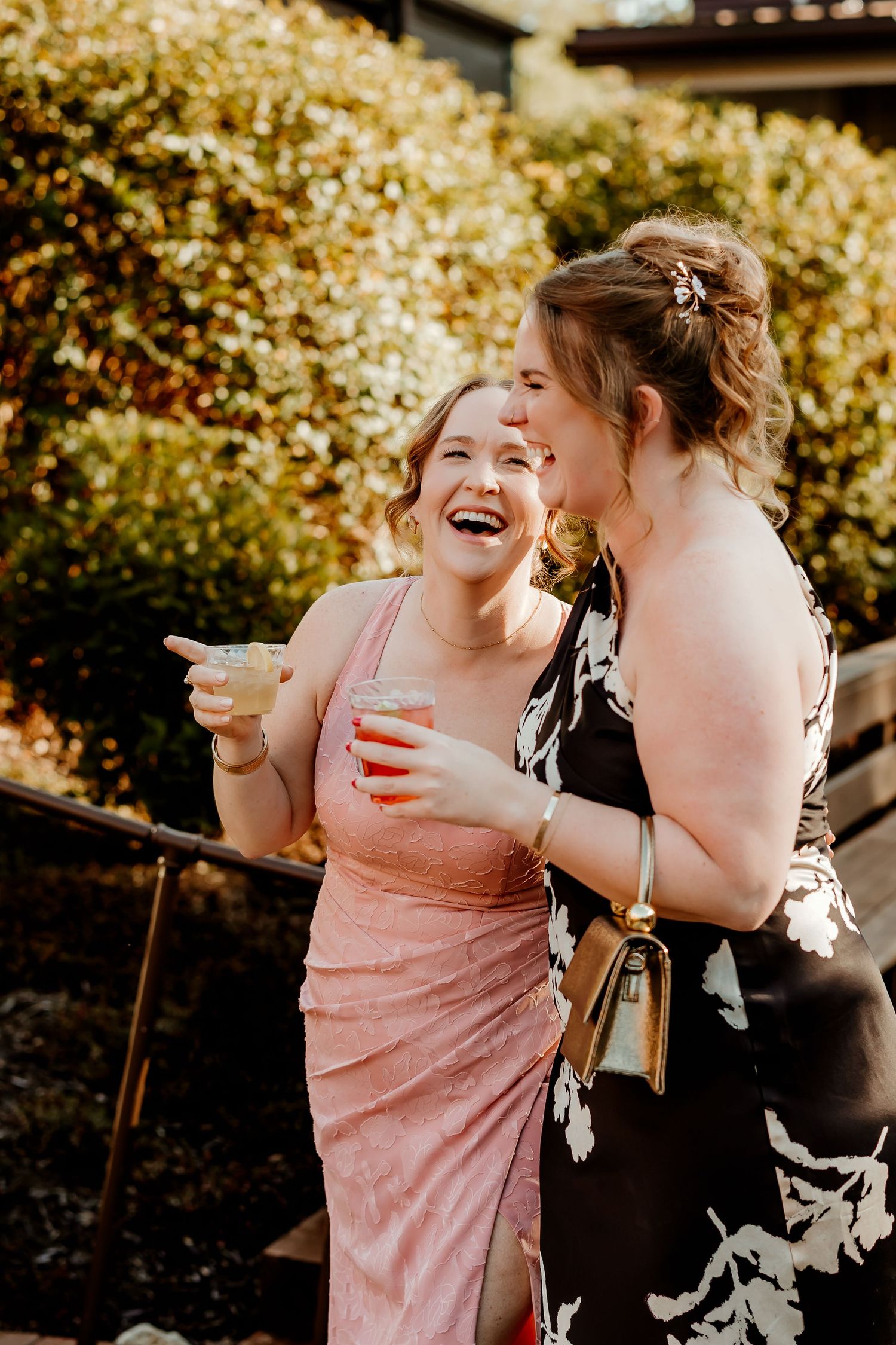 Two women laugh together while holding cocktails at an outdoor event, wearing a pink satin dress and a black floral dress.