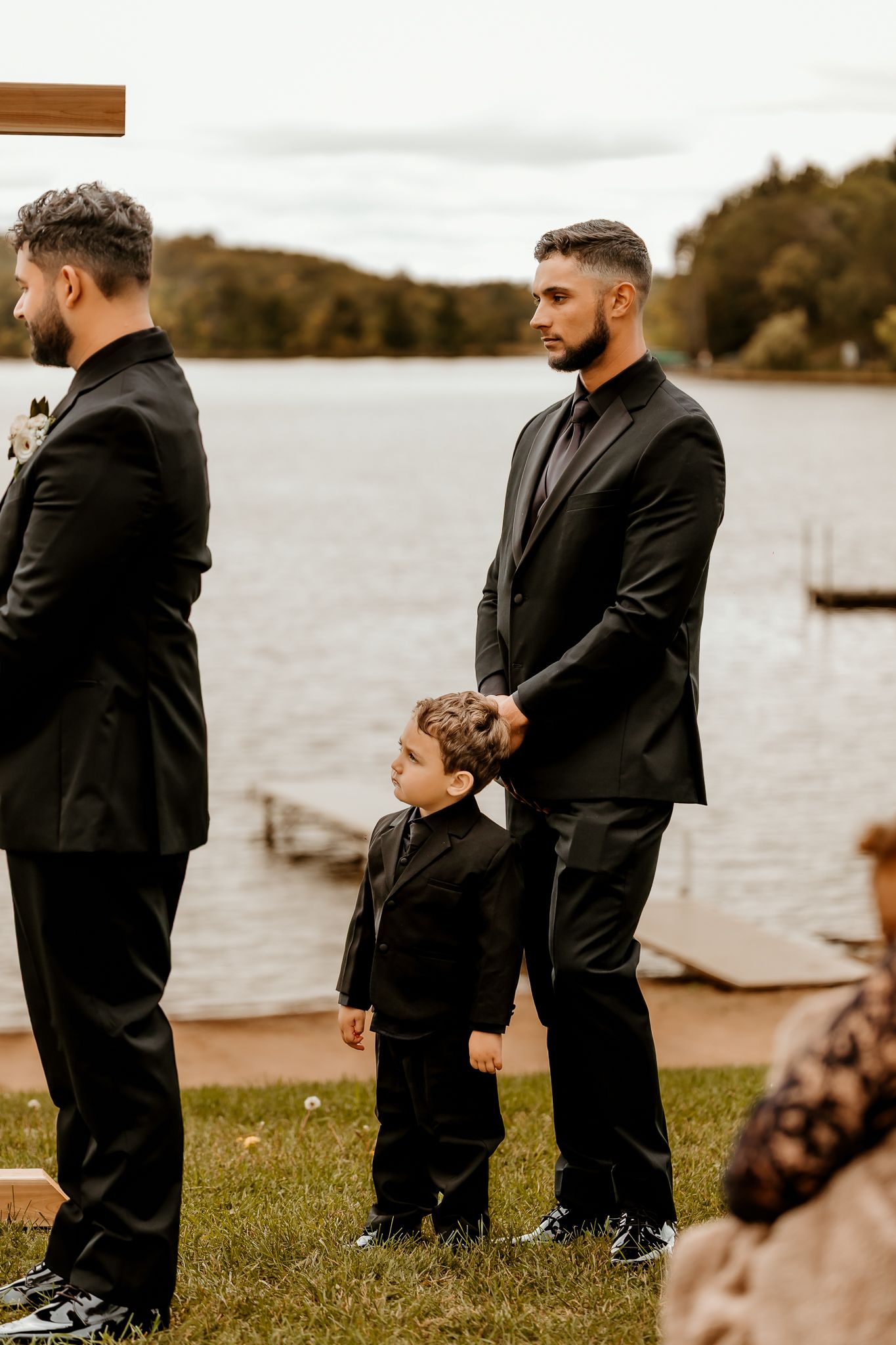 Two groomsmen in black suits and a young boy in a matching suit stand on a grassy lakeside lawn during an outdoor wedding ceremony.