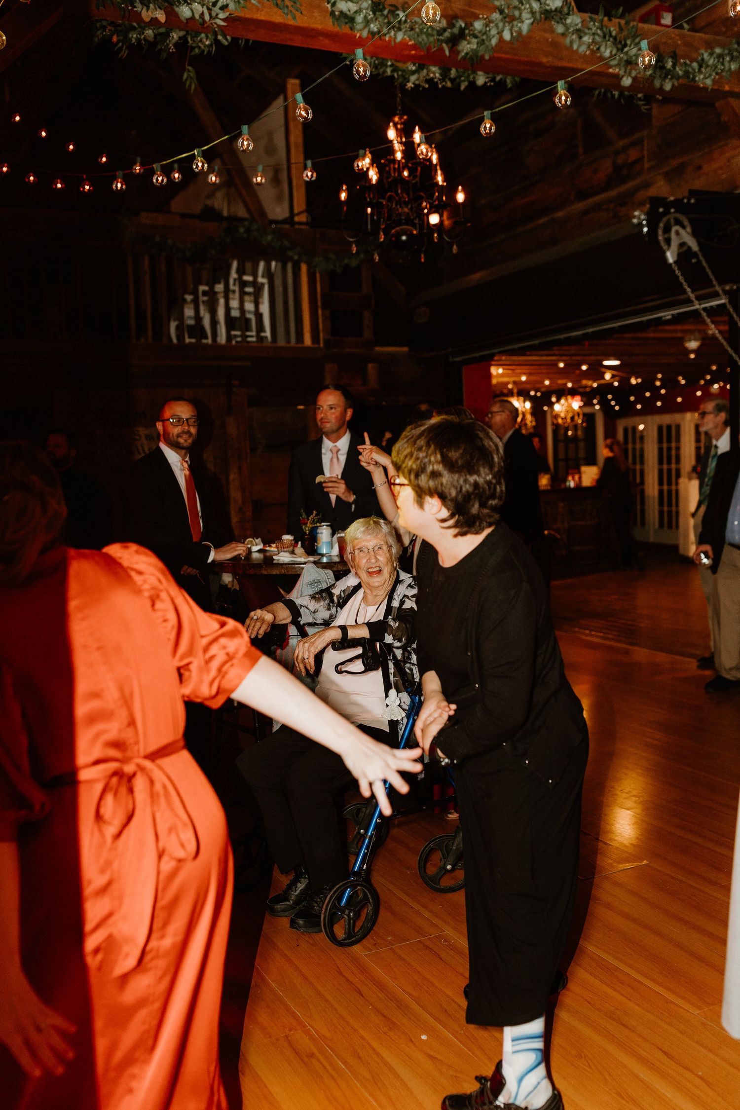 Guests dance on a wood floor at a rustic barn wedding reception, with an elderly woman in a wheelchair smiling beneath string lights.