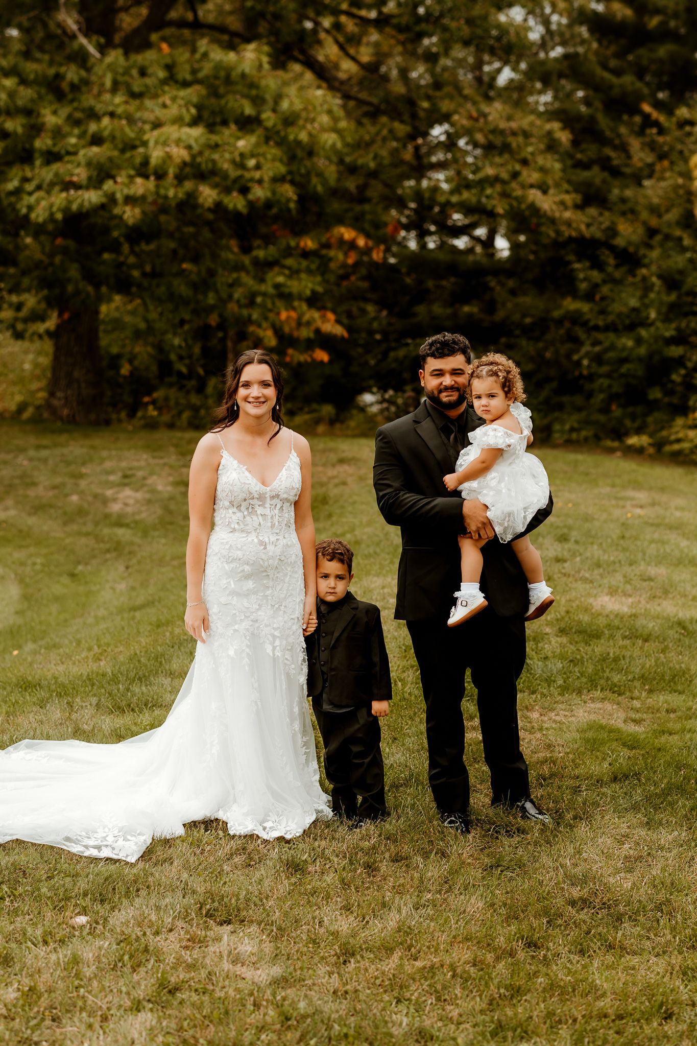 Bride in a lace wedding gown poses outdoors on a grassy lawn with a groom holding a toddler girl and a young boy in a black suit.