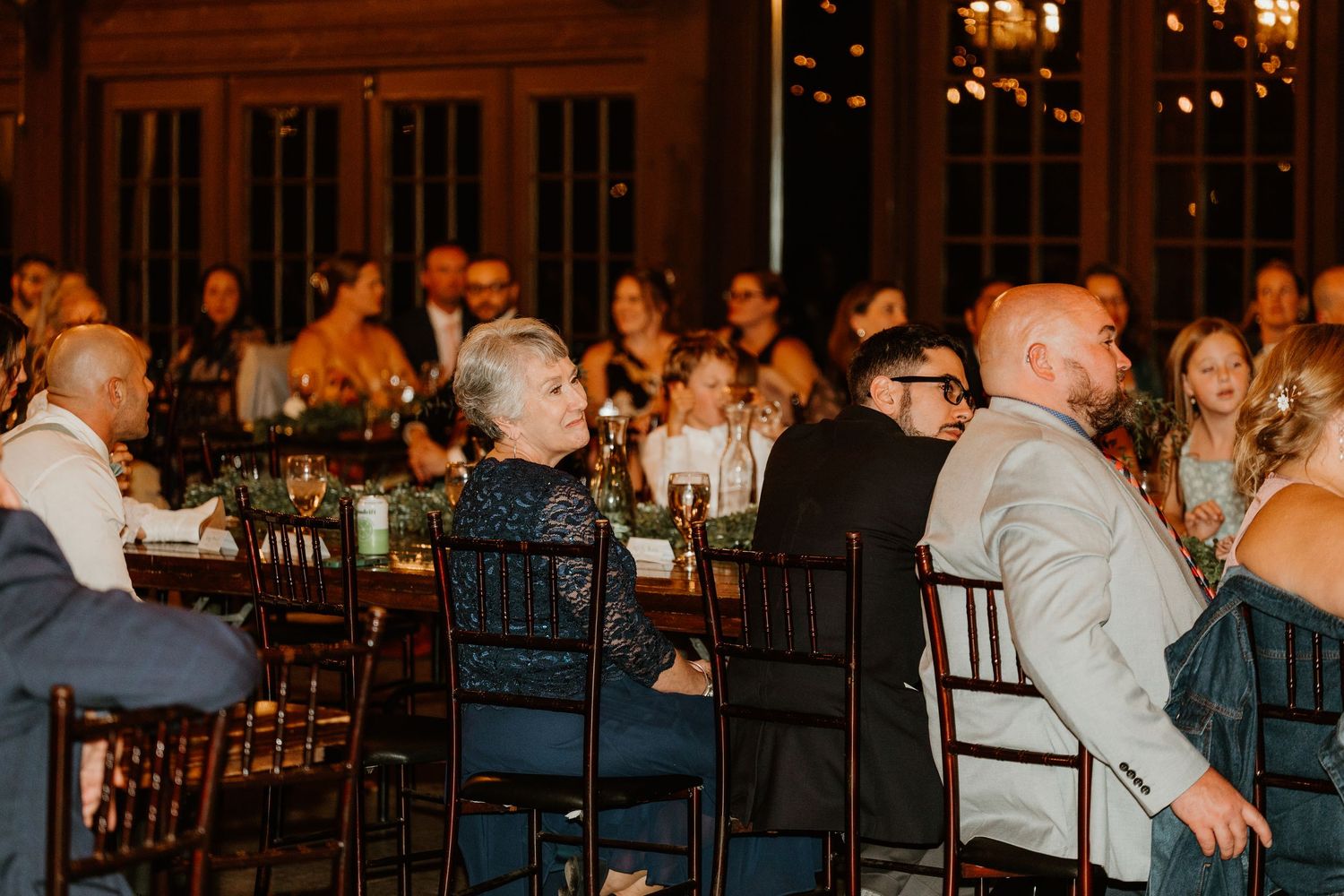 Wedding guests seated at dark wooden tables in an elegant, warmly lit reception hall, smiling and listening to a speech.