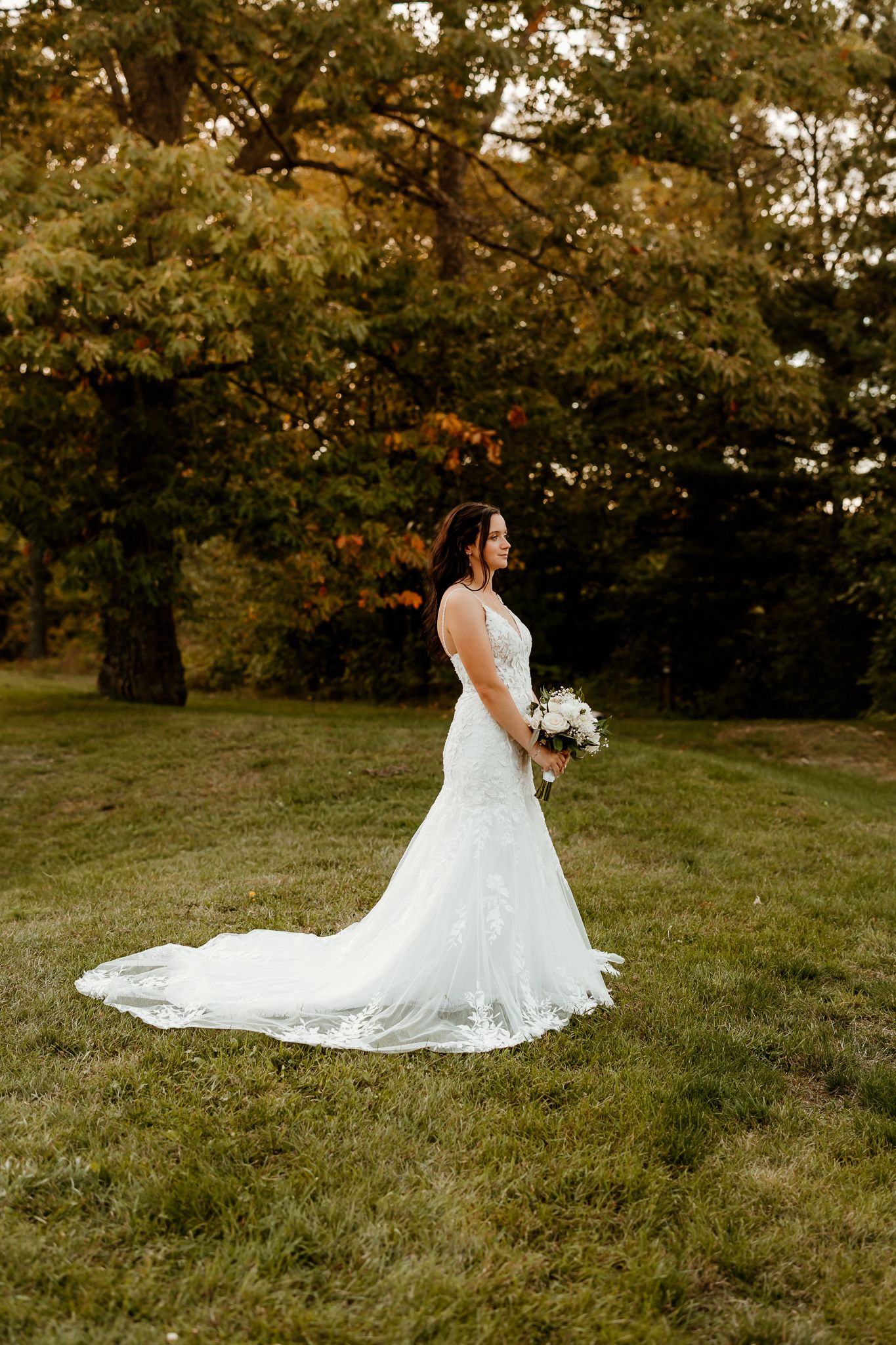 Bride in a lace wedding gown with a long train holds a white bouquet while standing on a grassy hill beside autumn trees.