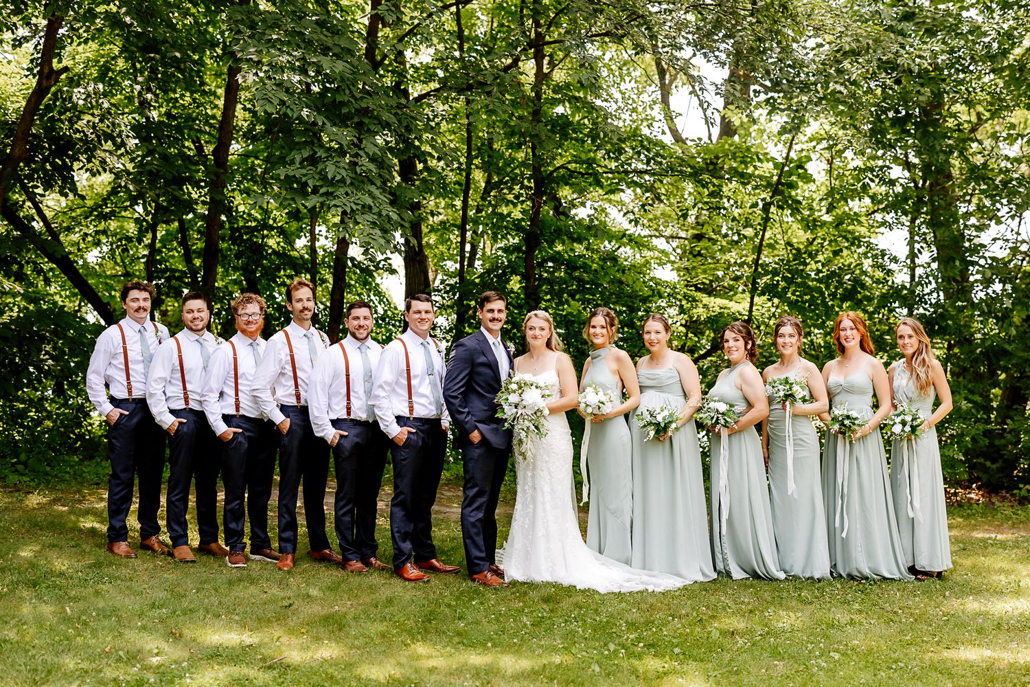 Wedding party poses outdoors in lush green setting, bridesmaids in sage gowns, groomsmen in navy suits with suspenders.