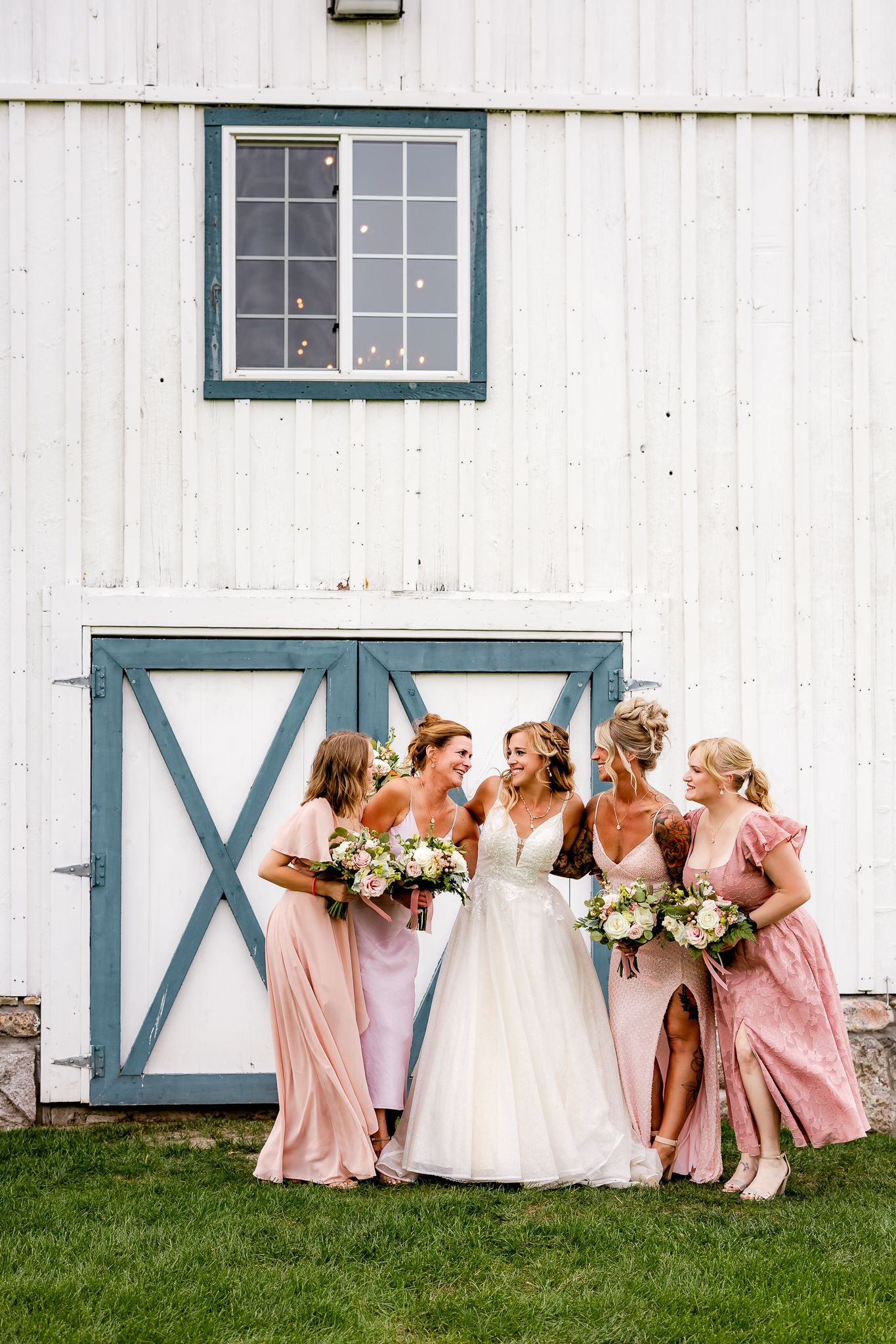 Bride in white gown with four bridesmaids in pink dresses holding bouquets, posing in front of a white barn.