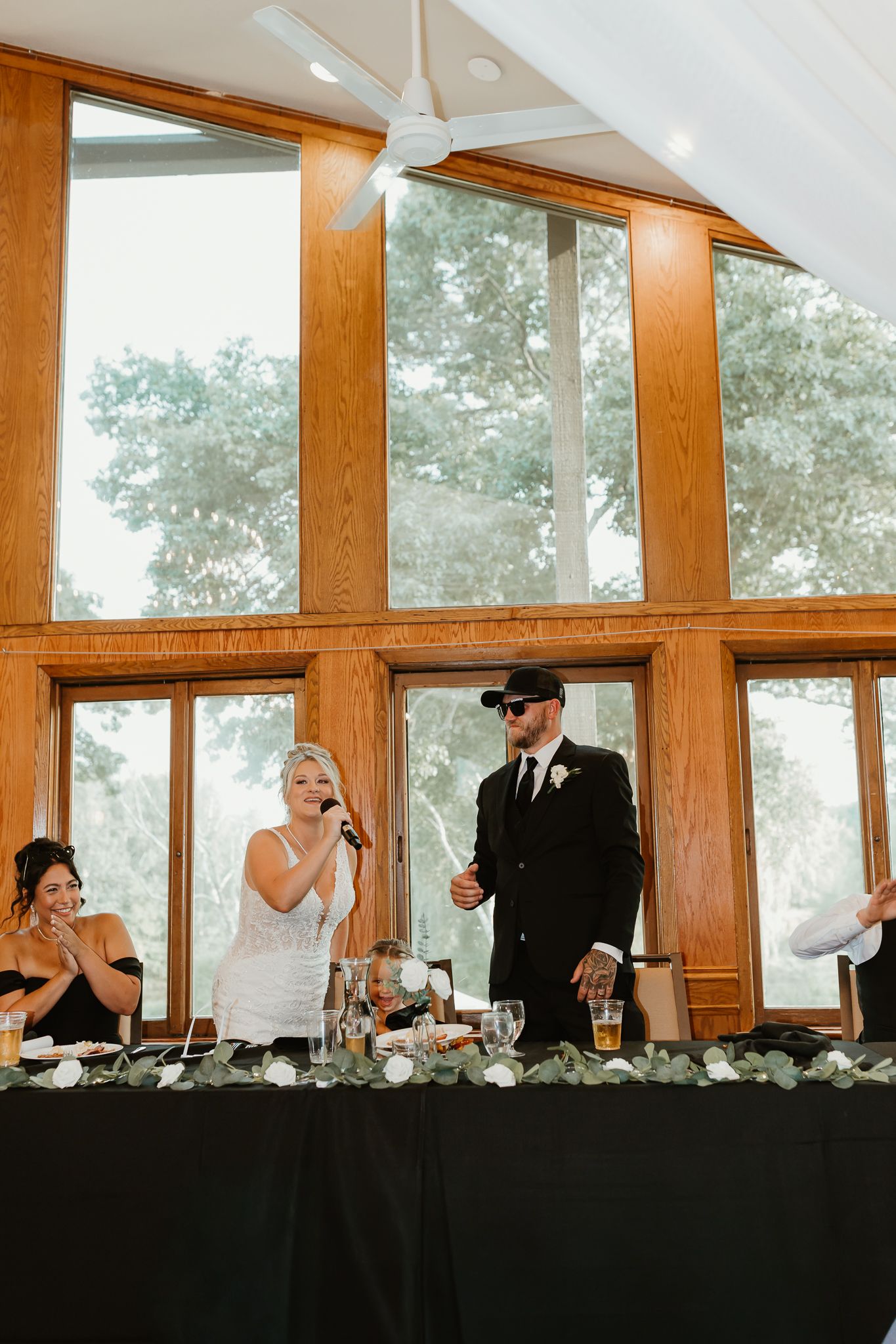 Bride and groom stand at wedding reception head table in elegant wood-paneled venue, bride speaking into microphone.