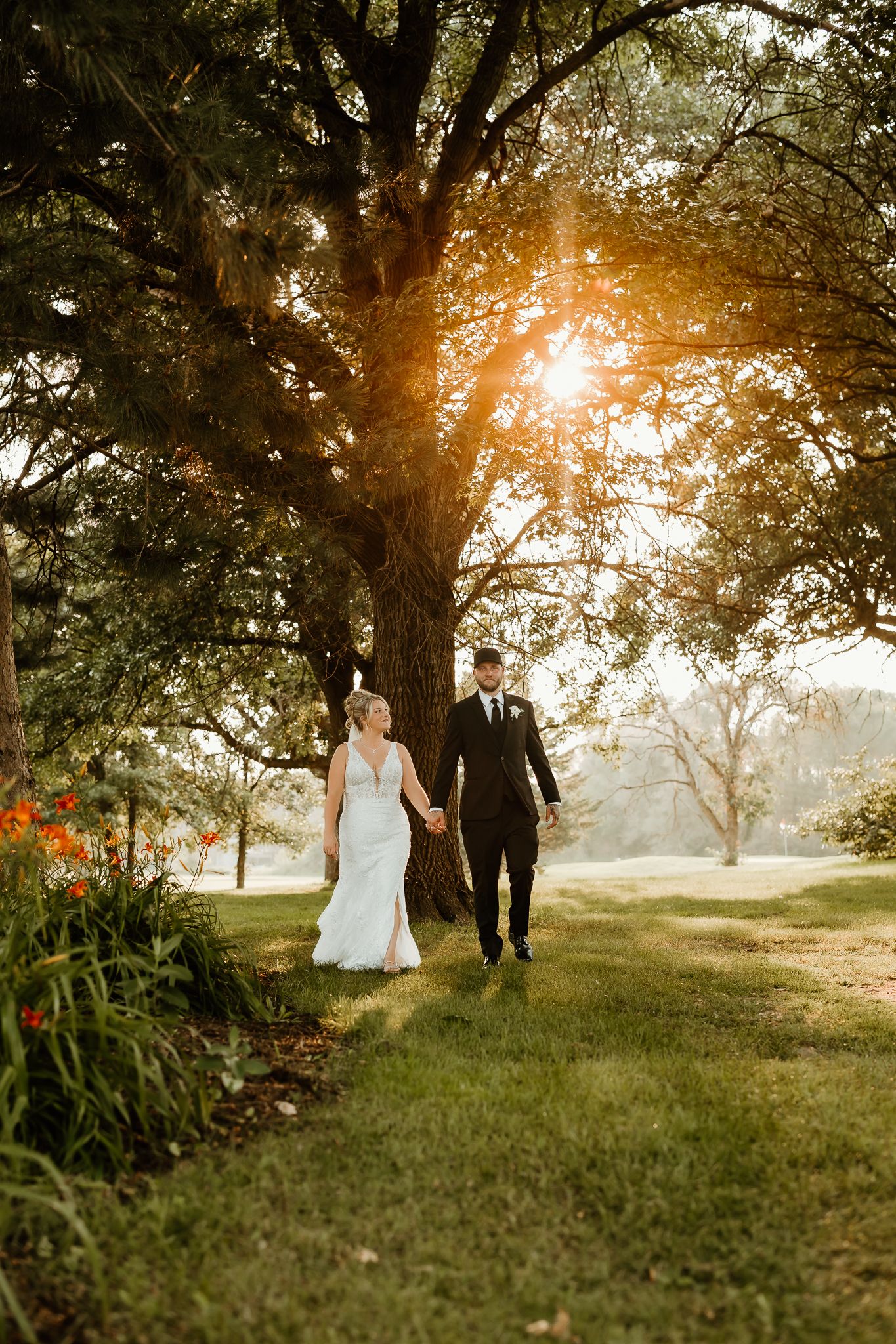A bride and groom walk hand in hand through a sunlit garden with a large tree glowing in golden hour light.