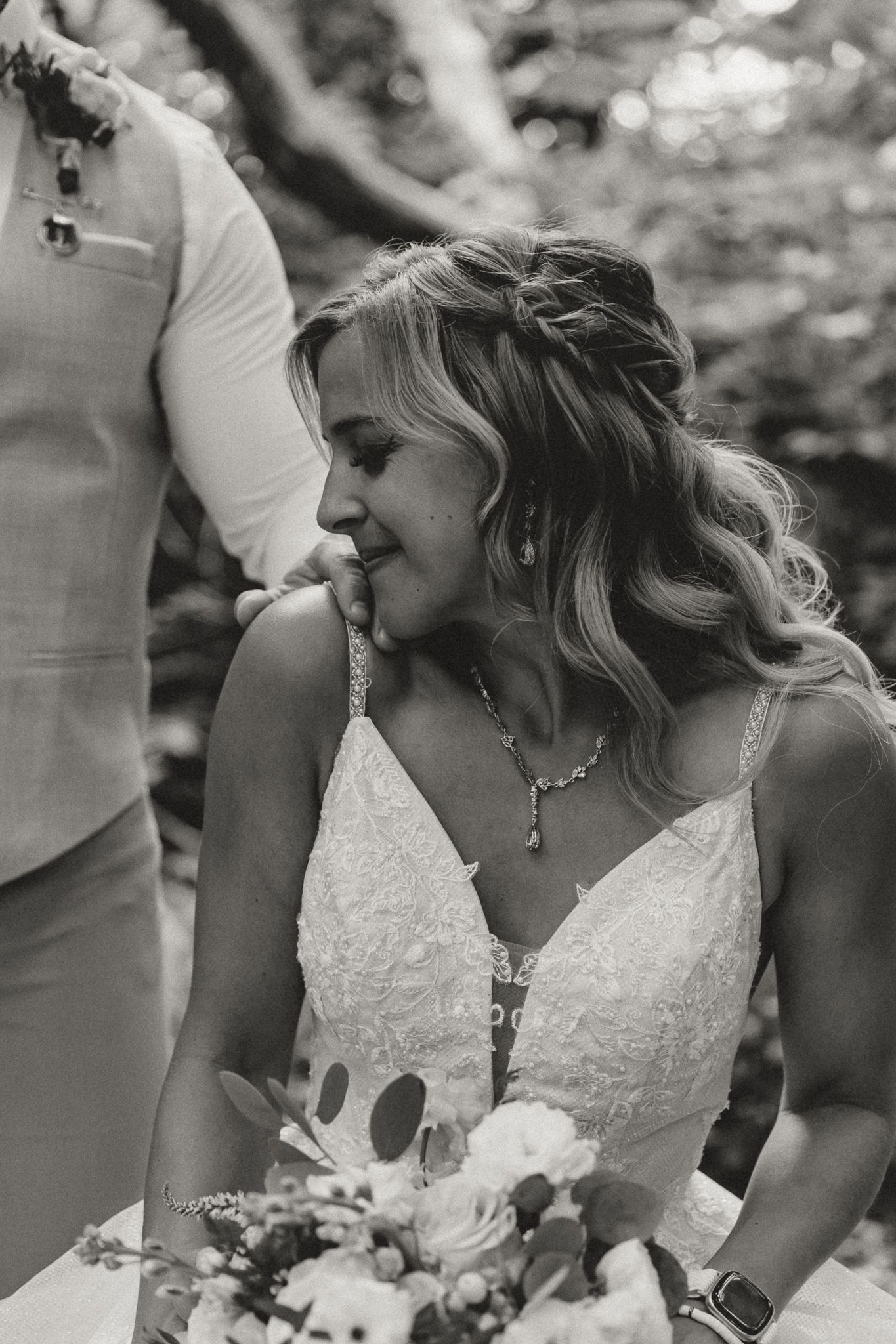 Black and white portrait of a smiling bride in lace gown holding a bouquet at an outdoor wedding ceremony.