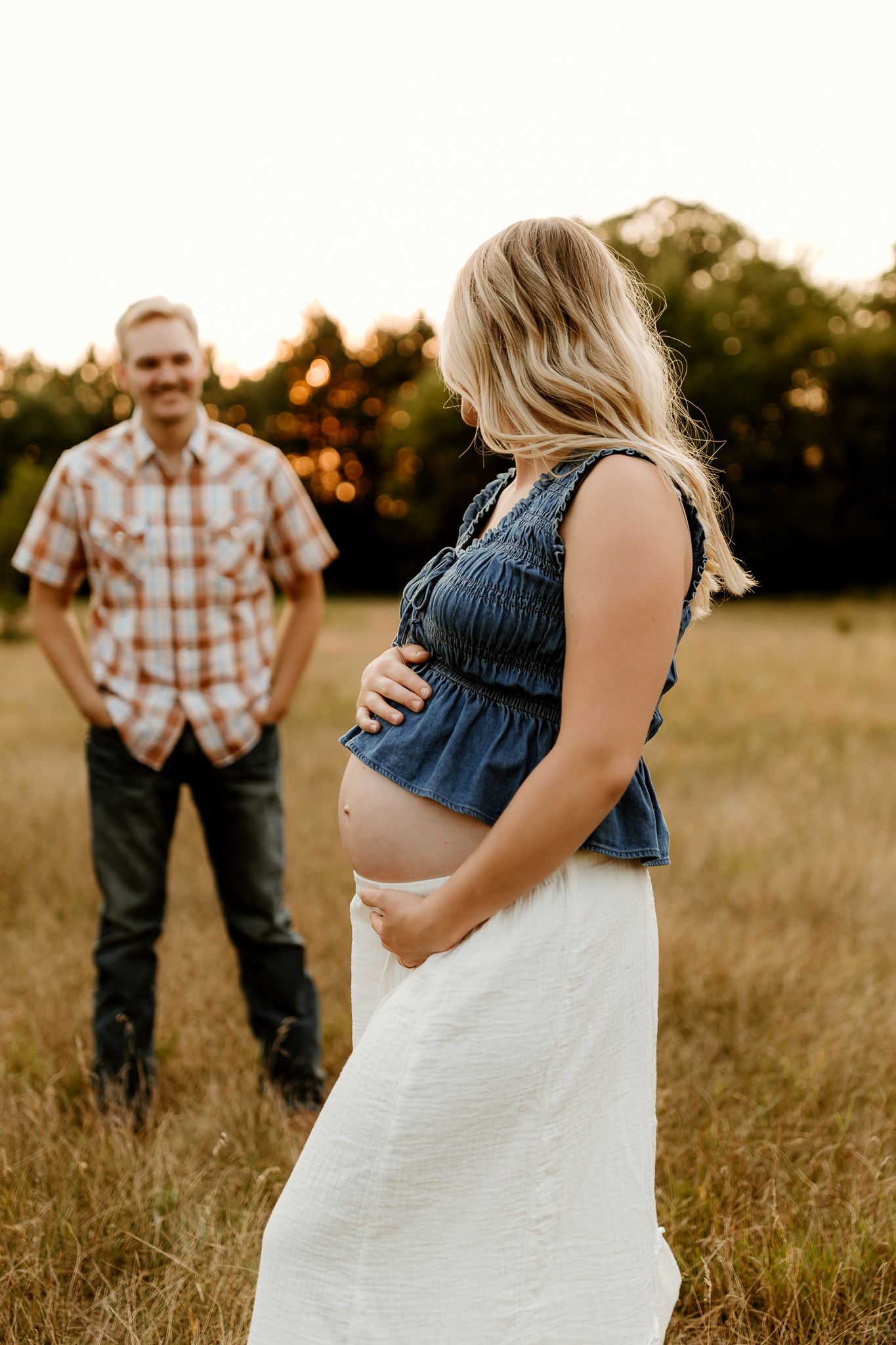 Pregnant woman in denim top and white skirt cradles baby bump in golden field while man in plaid shirt stands behind her.