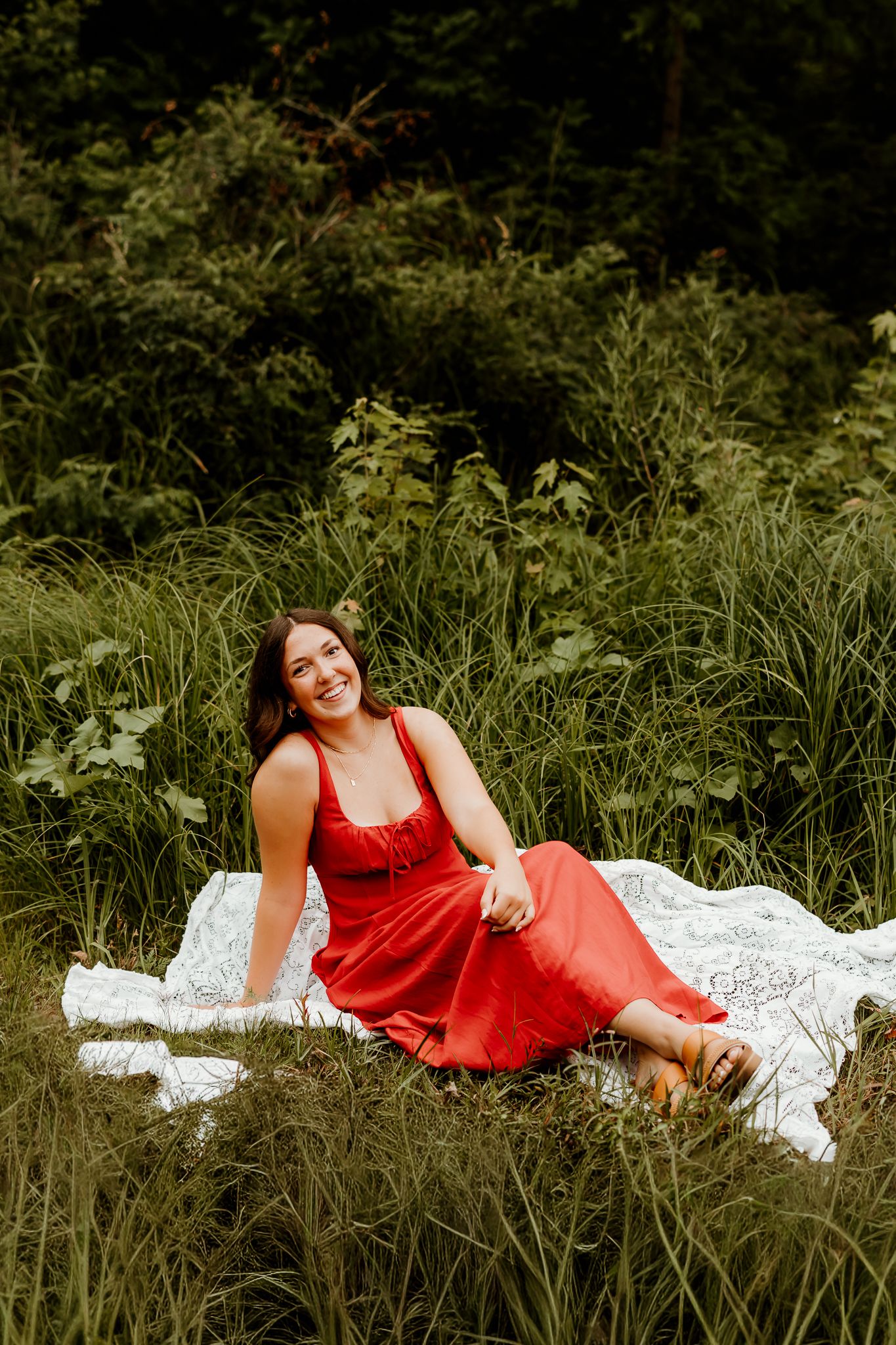 A smiling woman in a red dress sits on a white blanket surrounded by lush green grass and foliage outdoors.