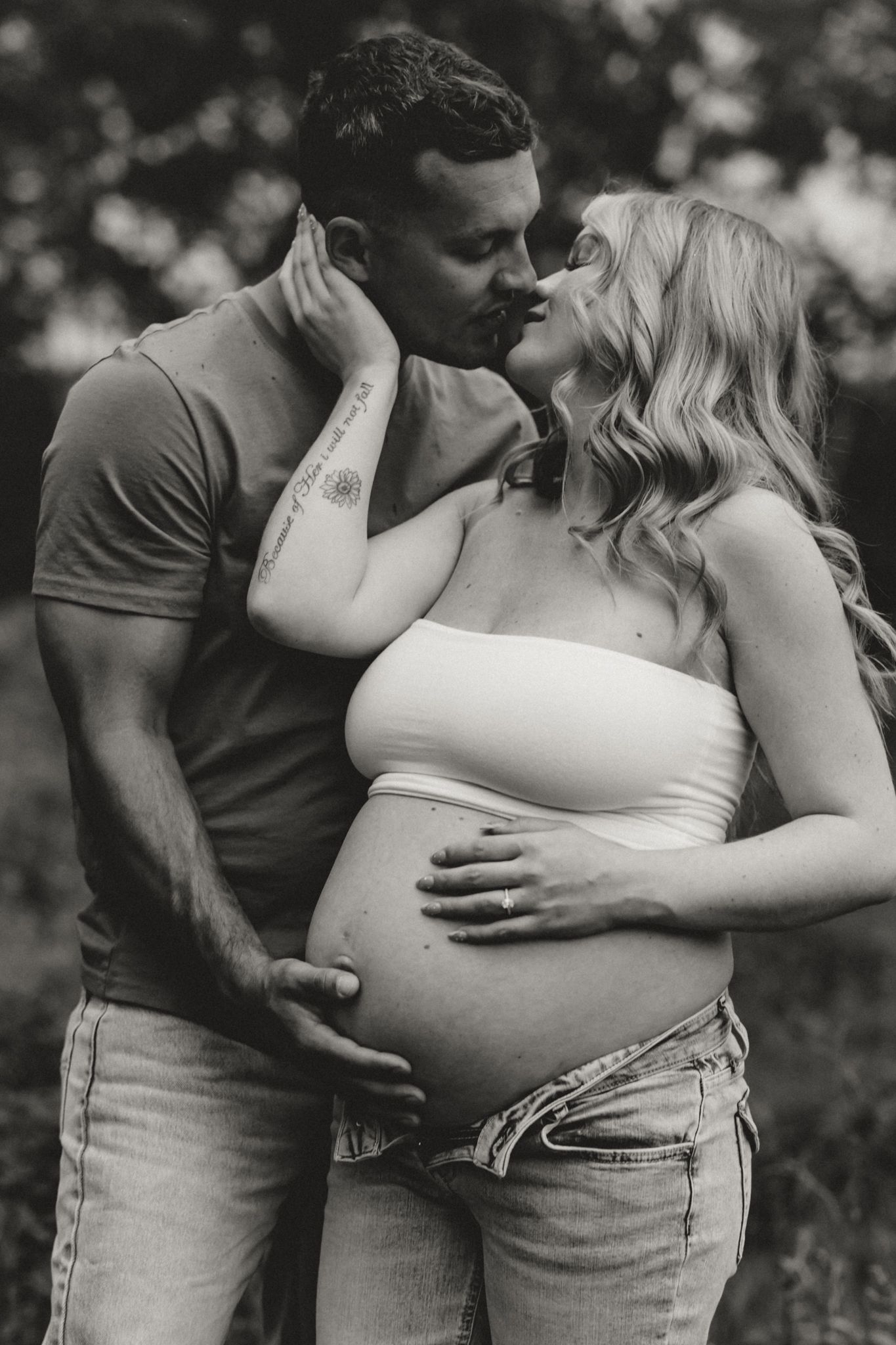 Black and white photo of a couple sharing a kiss while cradling a pregnant belly outdoors.