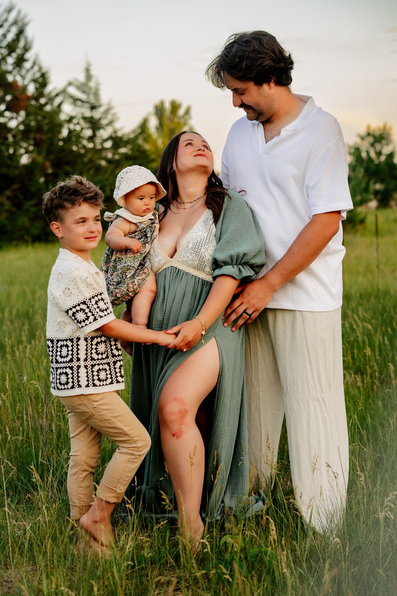 Family maternity photo in green field at sunset with pregnant mom in teal dress, dad, toddler, and young boy.