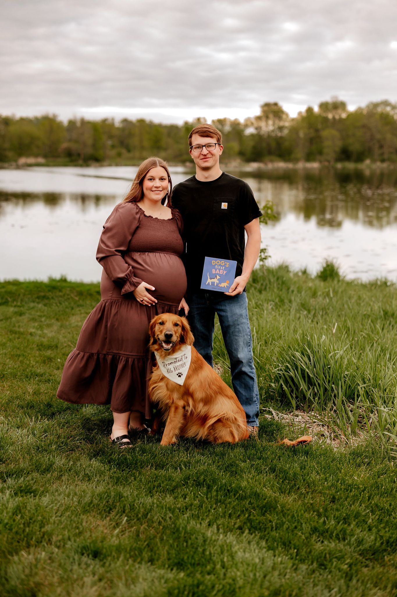 Expecting couple with golden retriever dog poses by lake holding baby book outdoors.