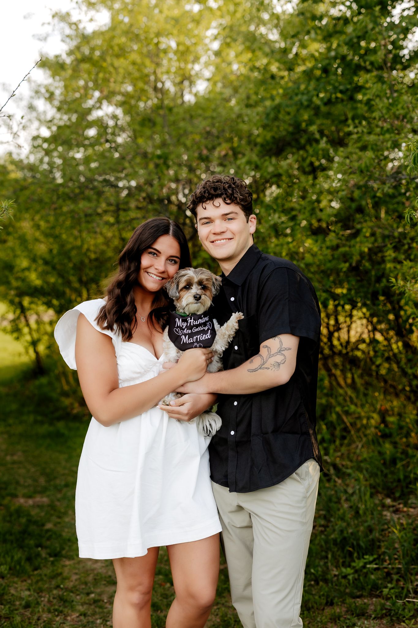 Smiling engaged couple holding a small dog with a 'Getting Married' bandana in a green outdoor setting.
