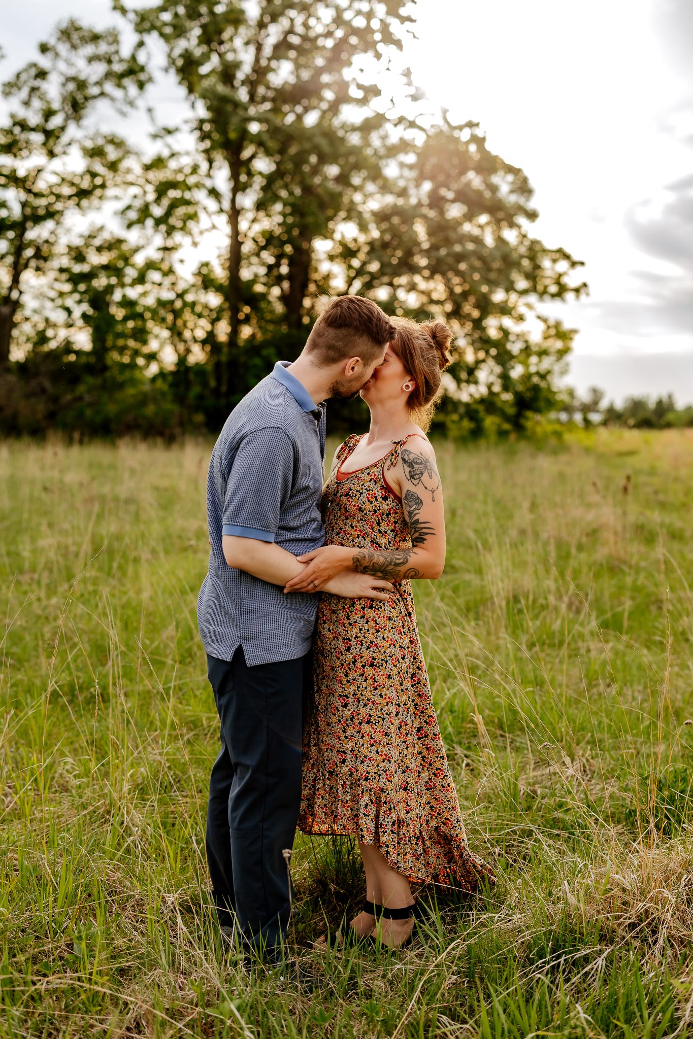 A couple shares a tender kiss in a golden sunlit field surrounded by lush green trees at sunset.