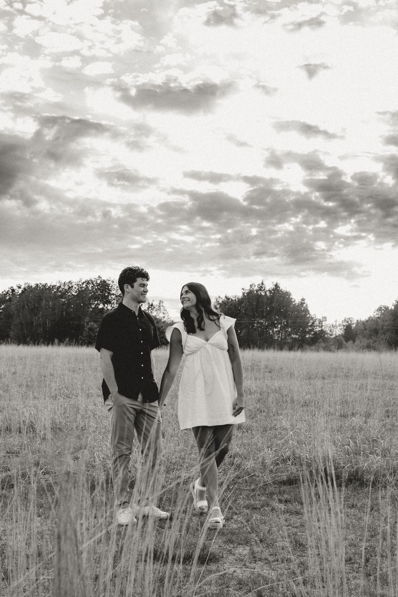 A black and white photo of a couple walking hand in hand through a grassy field under a cloudy sky.