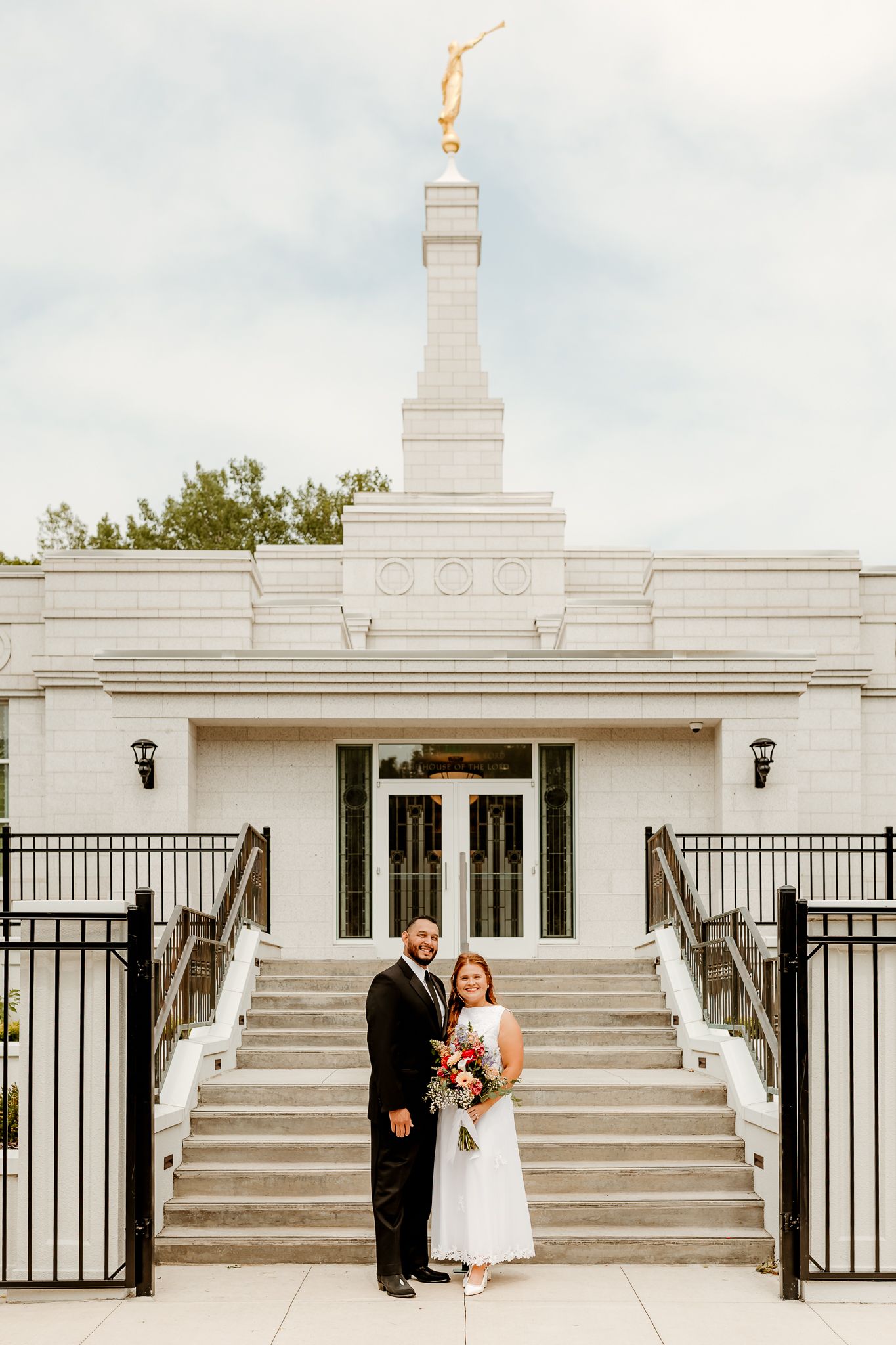 Newlywed couple posing in front of a white LDS temple with a golden spire on their wedding day.