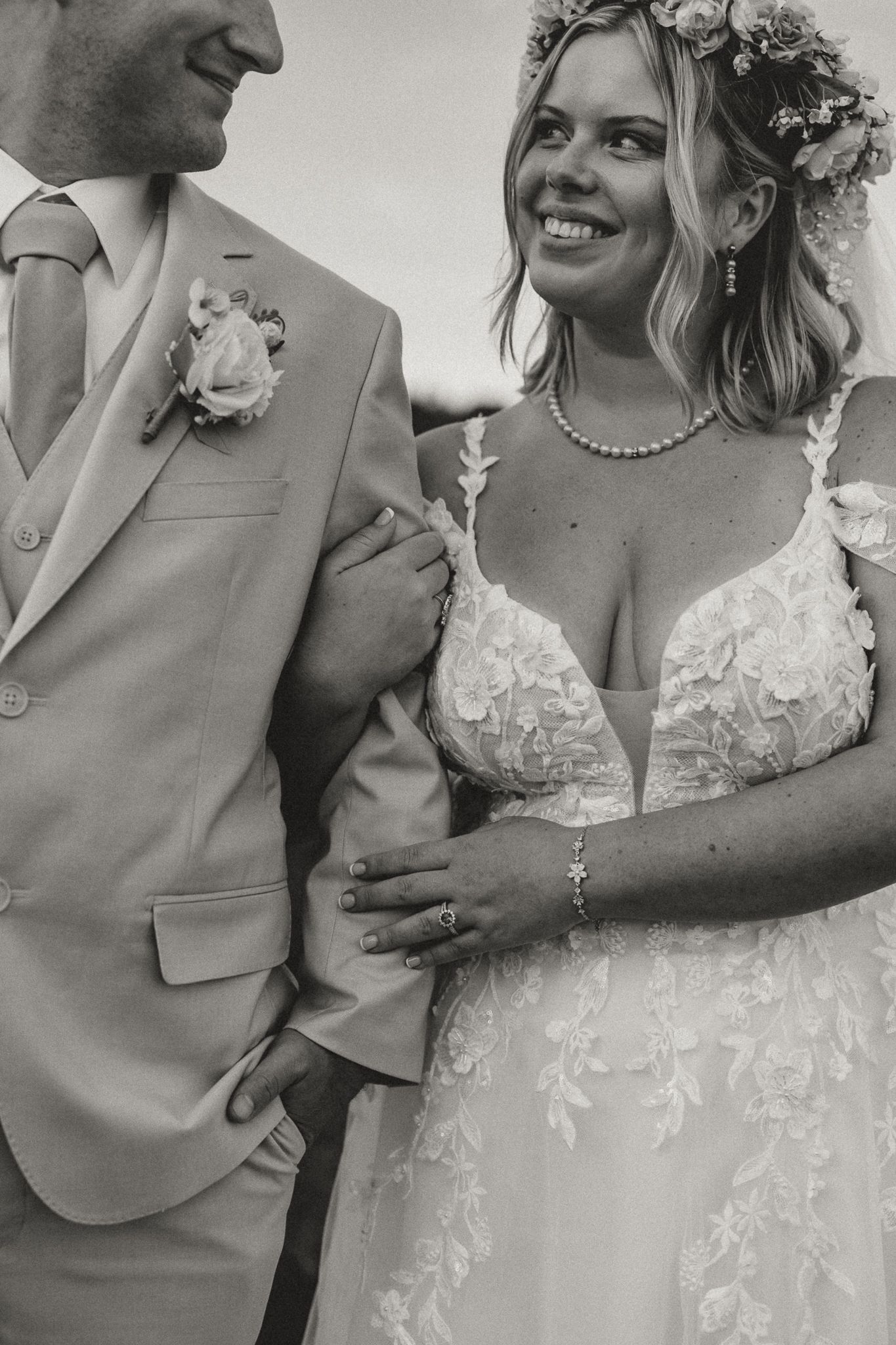 Black and white photo of a smiling bride in lace gown with flower crown standing beside groom in light suit.