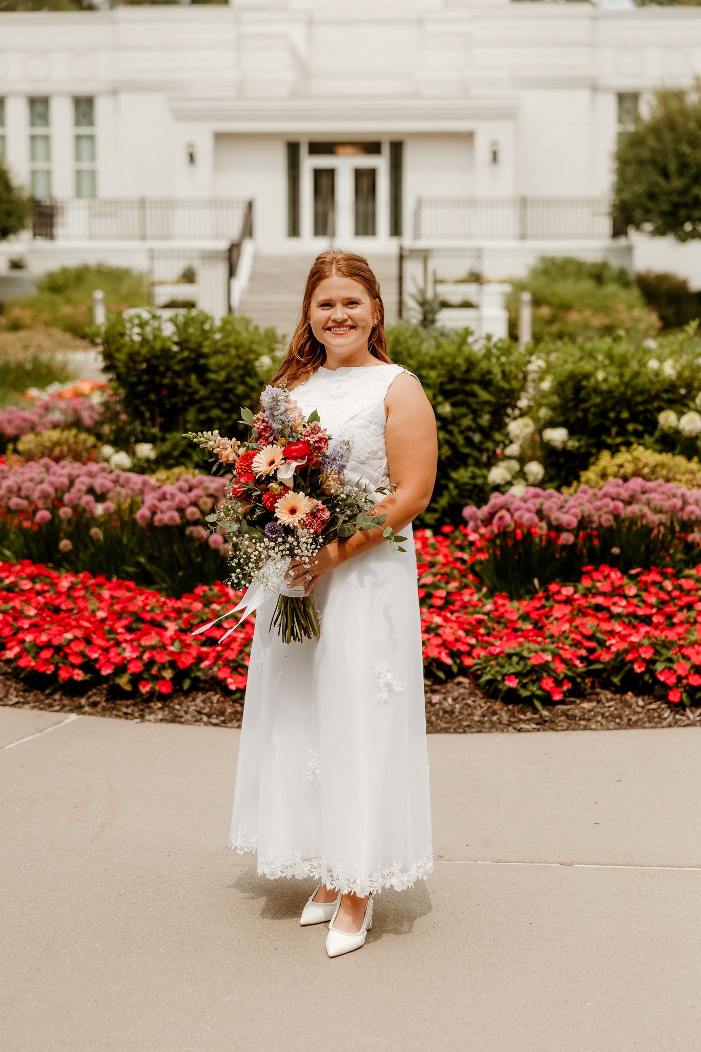 Bride in white lace dress holding colorful bouquet stands before red flower gardens at elegant venue.