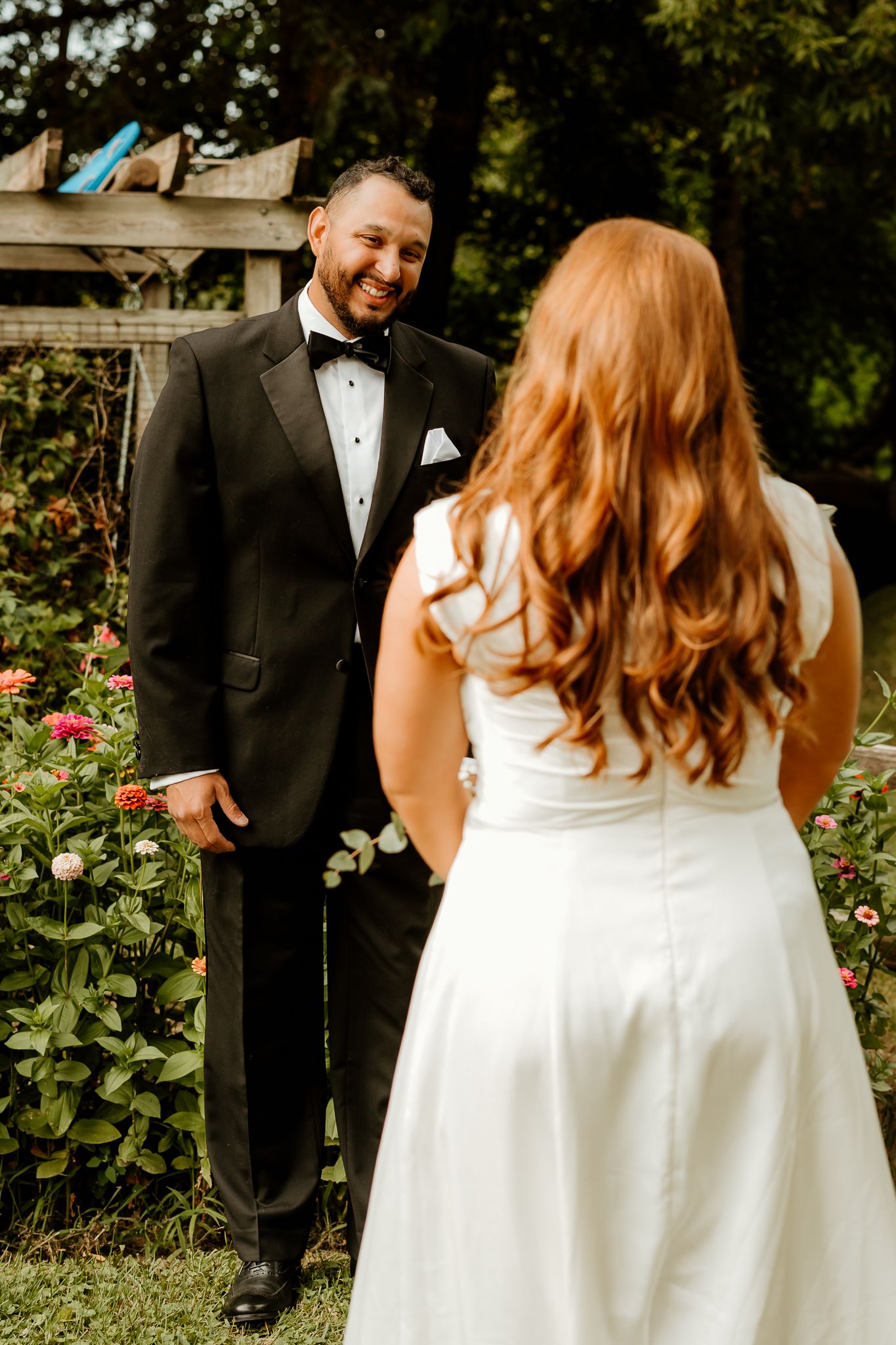 Groom in black tuxedo smiling at red-haired bride in white wedding dress during outdoor first look ceremony.