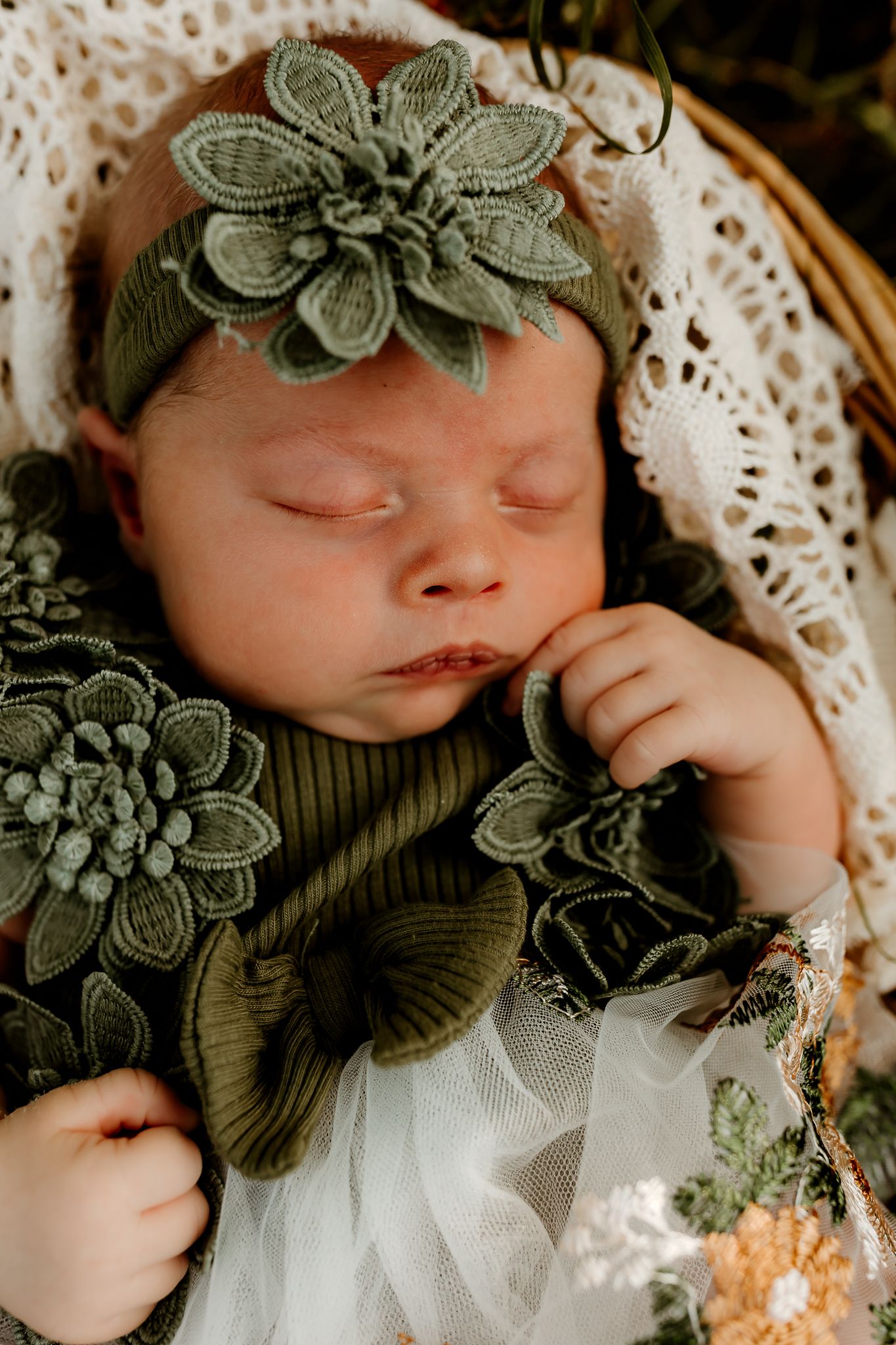 Sleeping newborn baby in olive green floral outfit and matching headband, nestled in a wicker basket with lace.