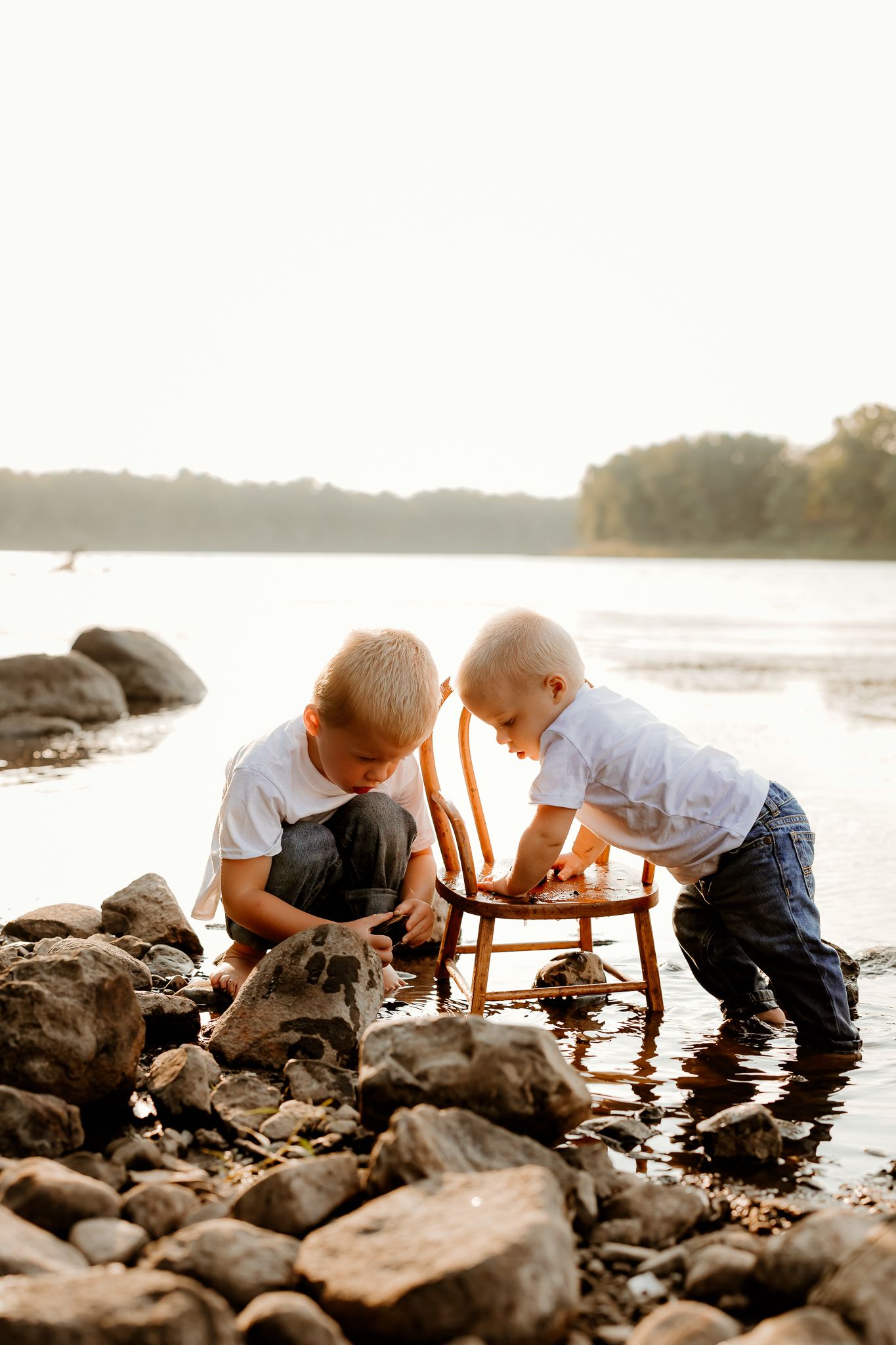 Two young boys playing with a wooden chair among rocks by a serene lake at golden hour sunset.