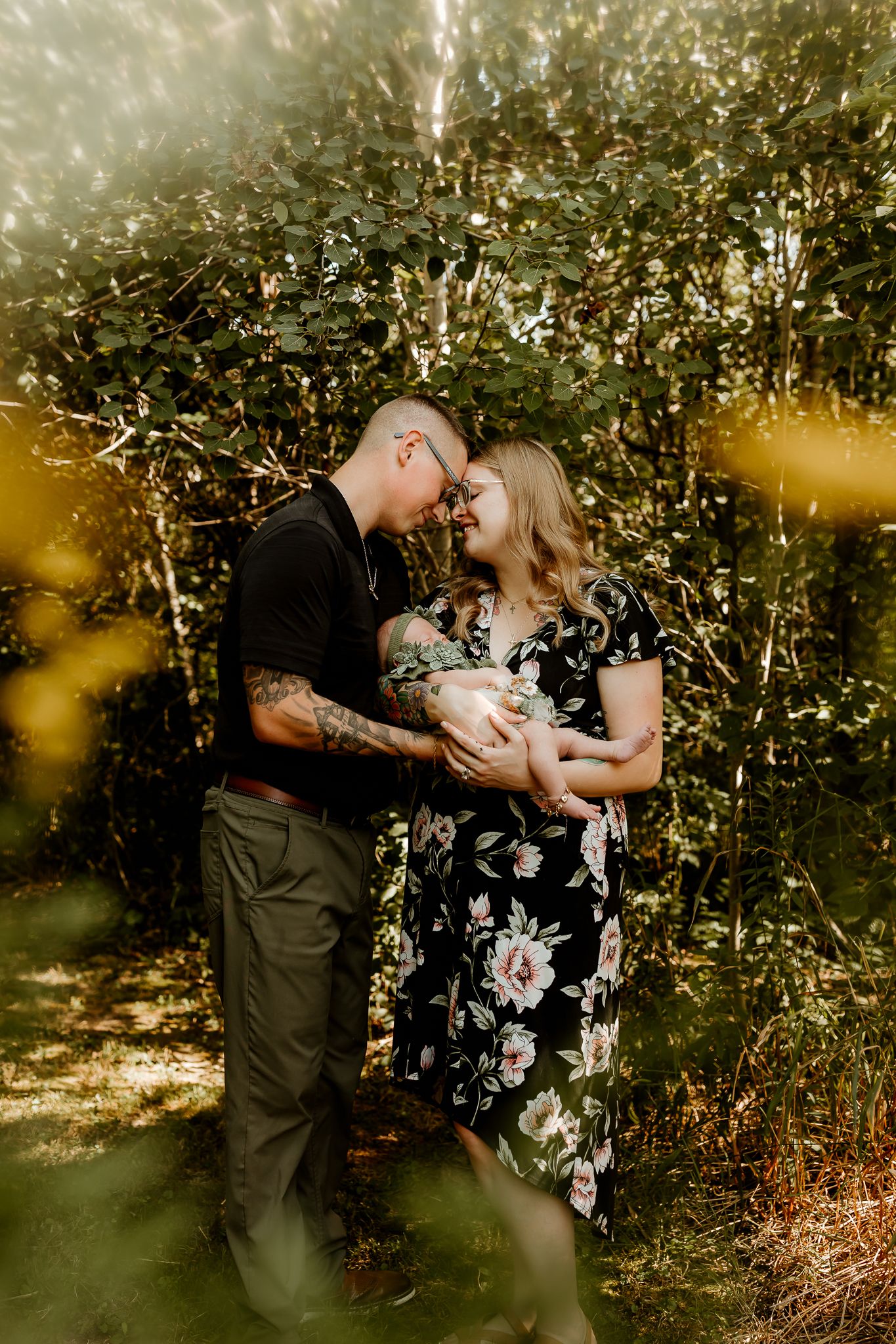 A couple tenderly touches foreheads while cradling their newborn baby outdoors surrounded by lush green foliage.
