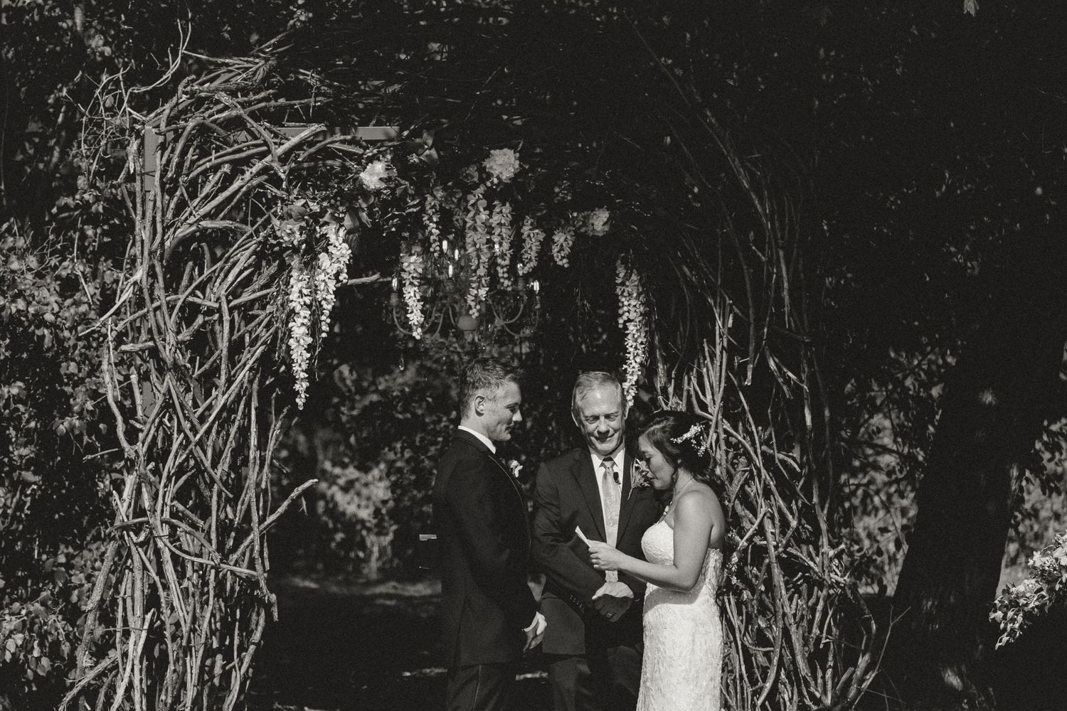 Black and white photo of a couple exchanging vows under a floral arch with an officiant at an outdoor wedding ceremony.