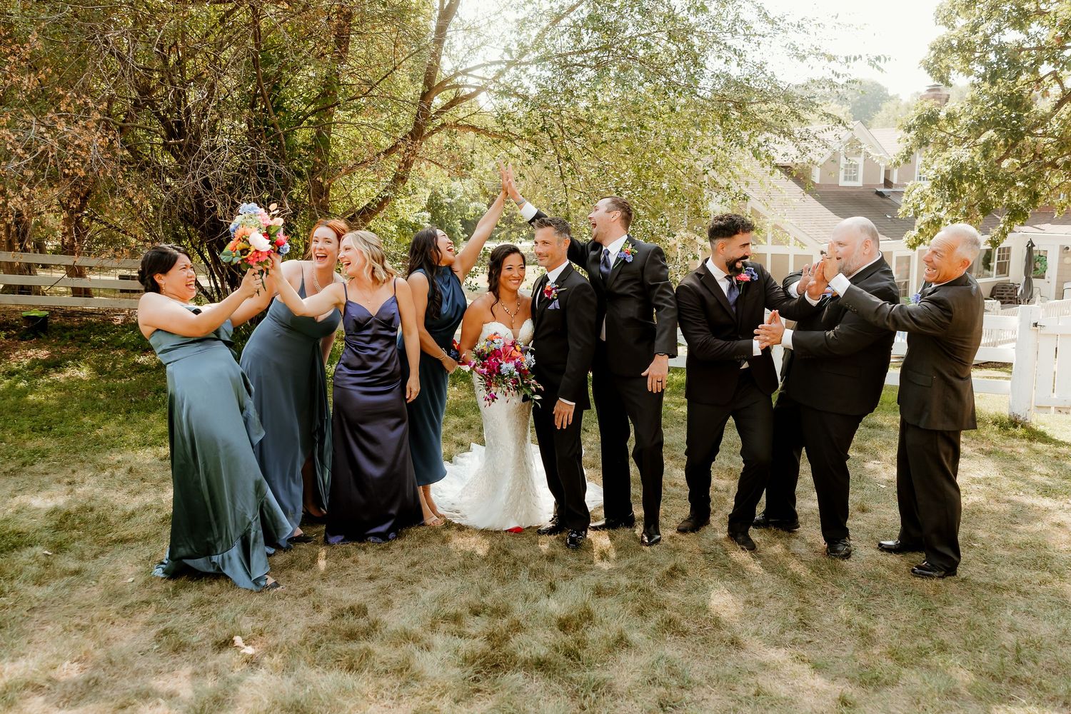 Wedding party celebrating outdoors with bride and groom surrounded by bridesmaids in blue gowns and groomsmen in black suits.