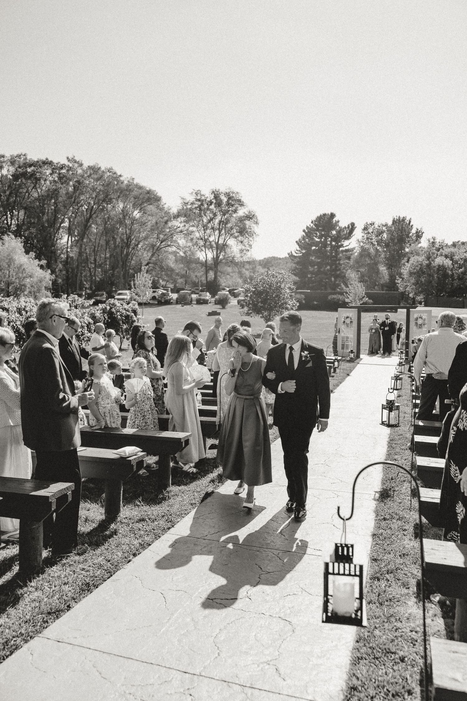 Black and white photo of a couple walking down the outdoor wedding aisle lined with wooden benches and lanterns.