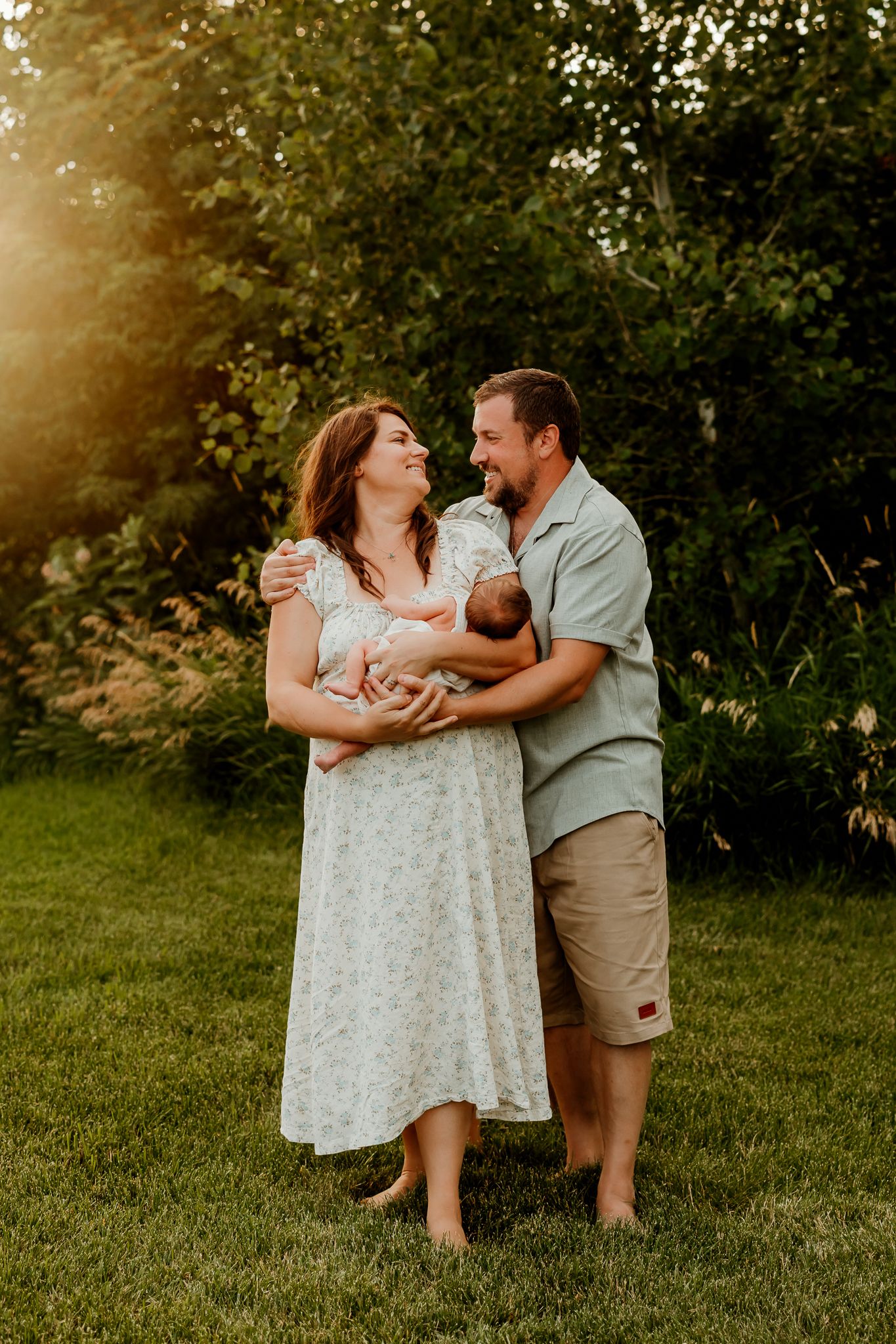A couple smiles at each other while holding their newborn baby outdoors in golden hour sunlight.