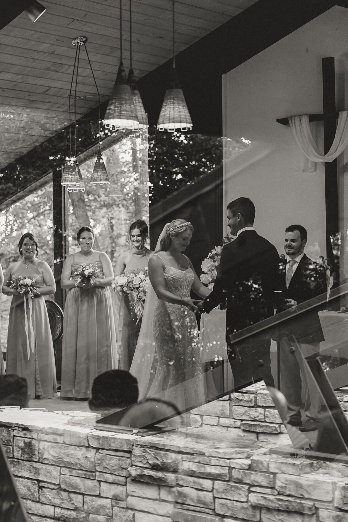 Black and white wedding ceremony photo with bride and groom holding hands, bridesmaids holding bouquets, at elegant venue.