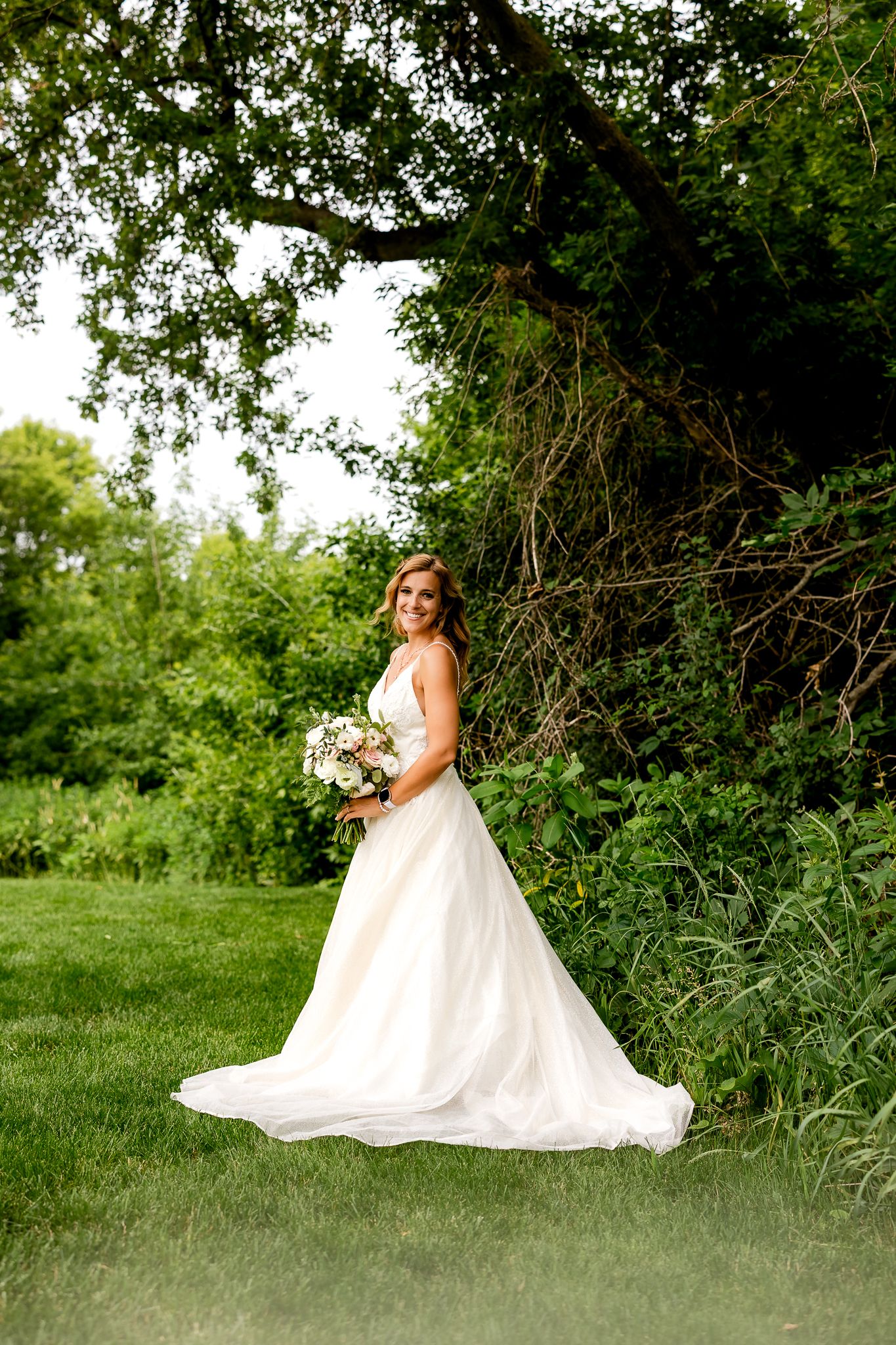 Bride in white ball gown holding bouquet stands on green lawn under large tree at outdoor wedding venue.