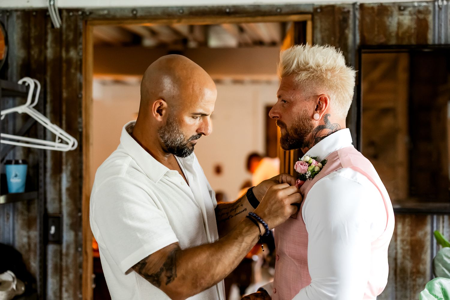 A bald man pins a boutonniere on a blonde groom in a pink suit during wedding preparations indoors.