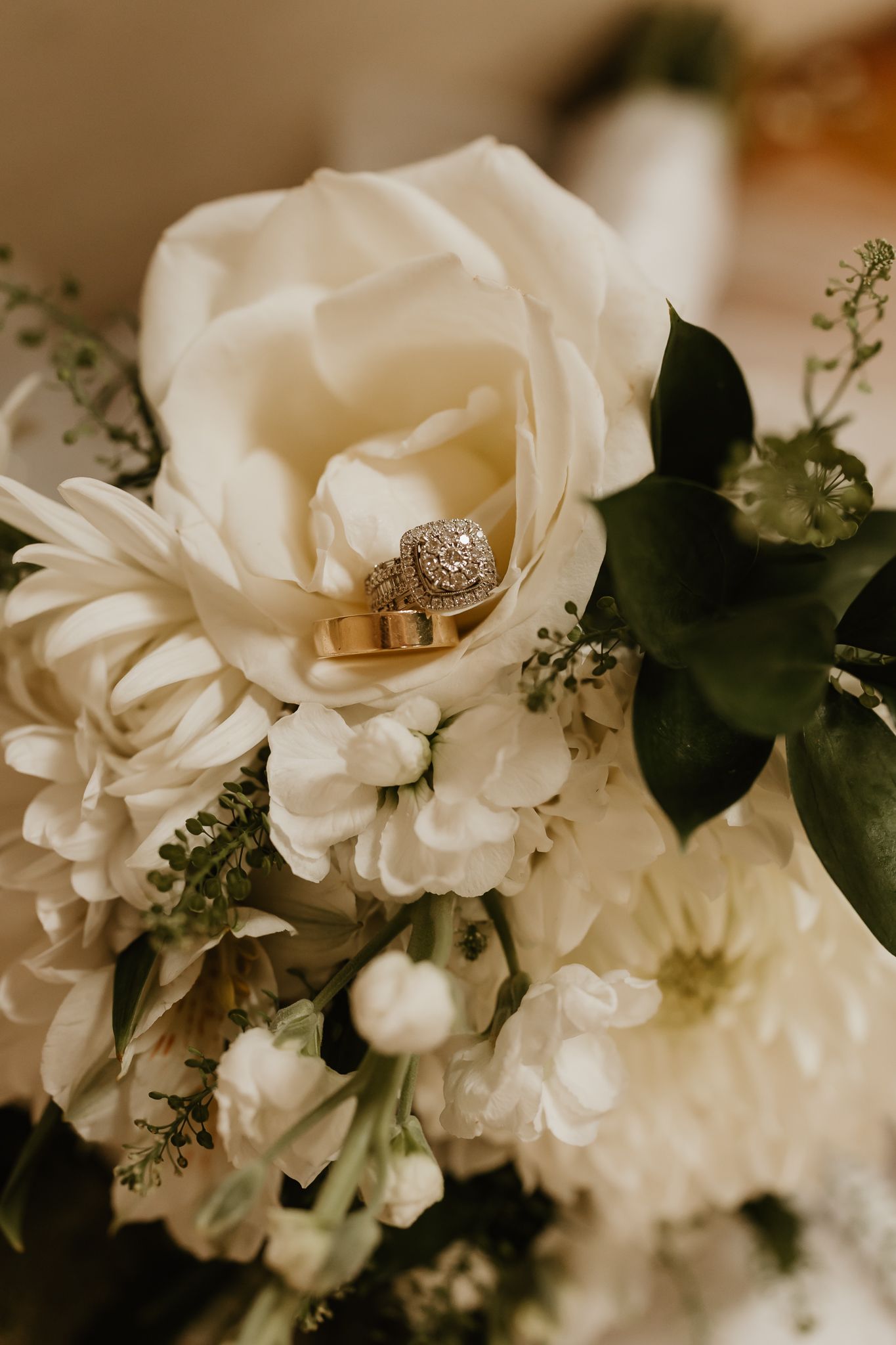 Wedding rings resting inside a white rose surrounded by white dahlias and greenery in a bridal bouquet.